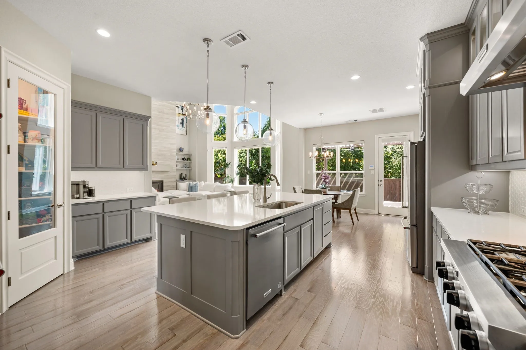 Chef's kitchen featuring a large center island with waterfall quartz countertop, custom gray cabinetry, stainless steel appliances including dual cooktops, pendant lighting, and abundant natural light from multiple windows overlooking the backyard