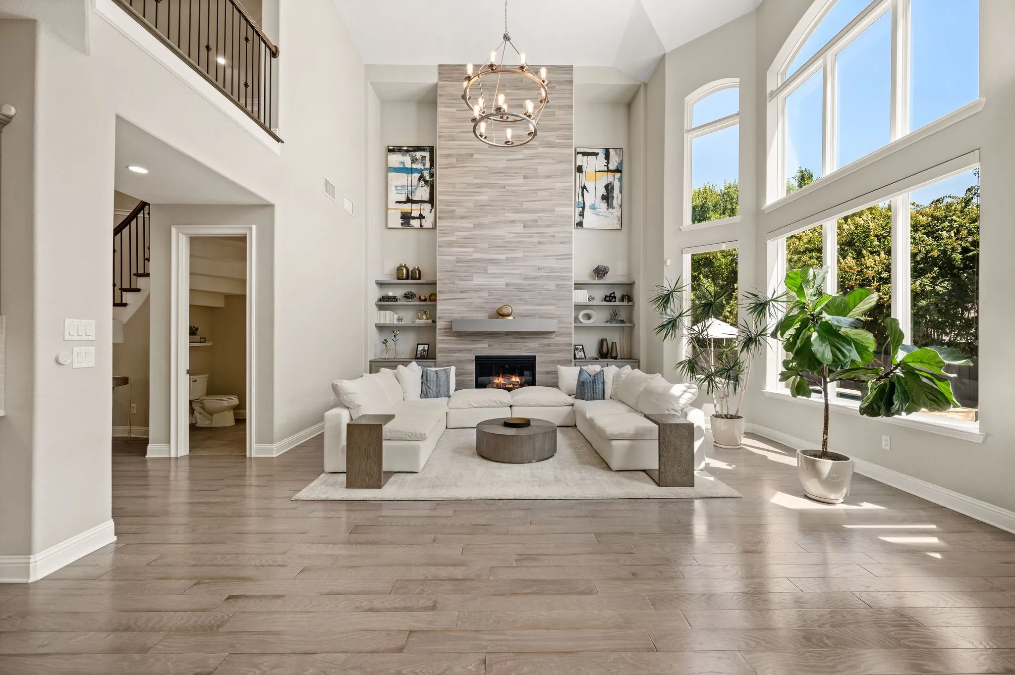 Two-story living room with soaring ceilings, floor-to-ceiling windows framing natural light and outdoor scenery, wide-plank wood-finish flooring throughout, a linear fireplace with stacked stone surround and built-in shelving, and a modern chandelier suspended from the ceiling