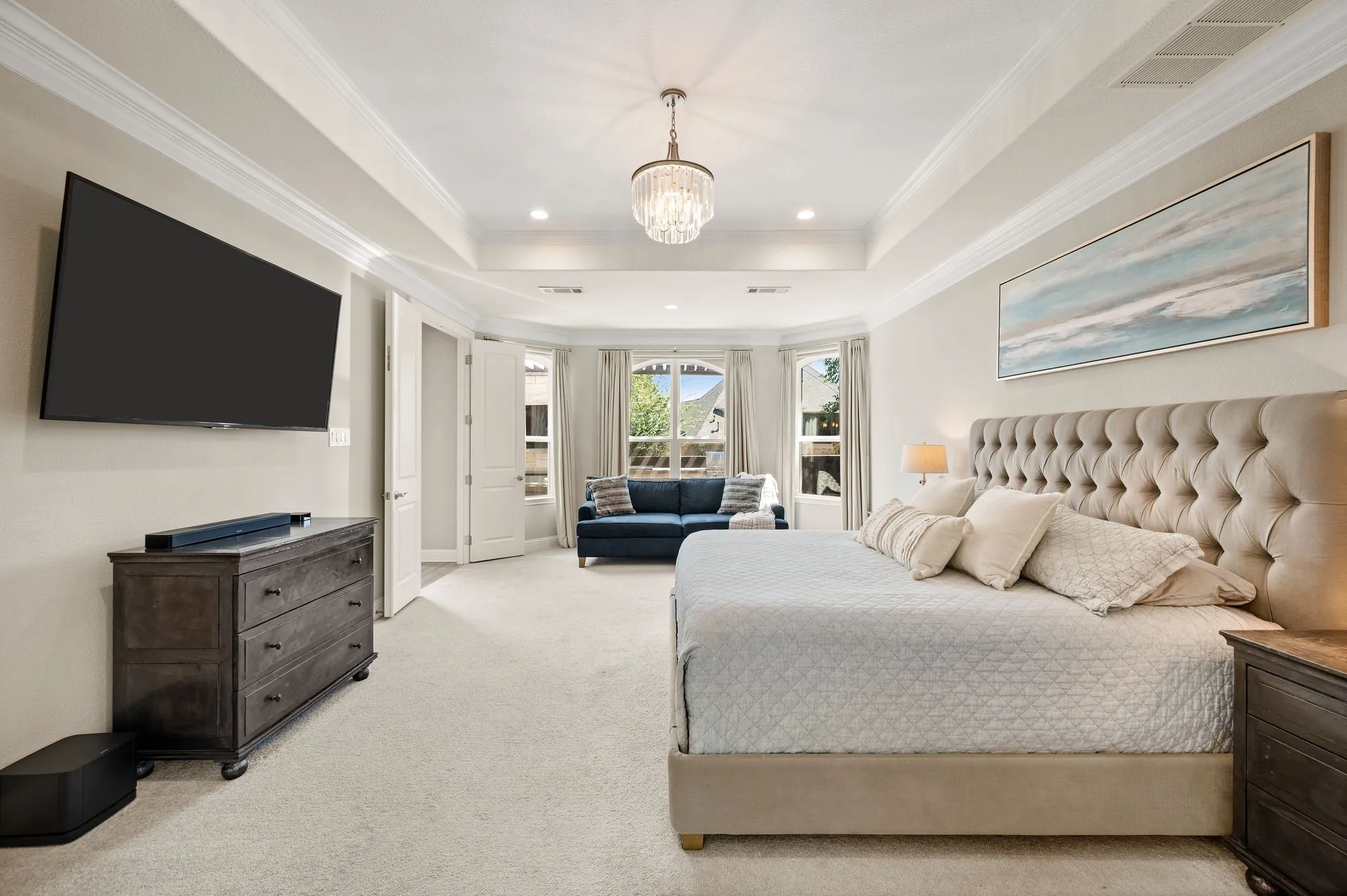 Luxurious primary suite featuring a coffered ceiling with integrated lighting, dual nightstands with wood-finish construction, an upholstered headboard with button detailing, and plush carpeting throughout