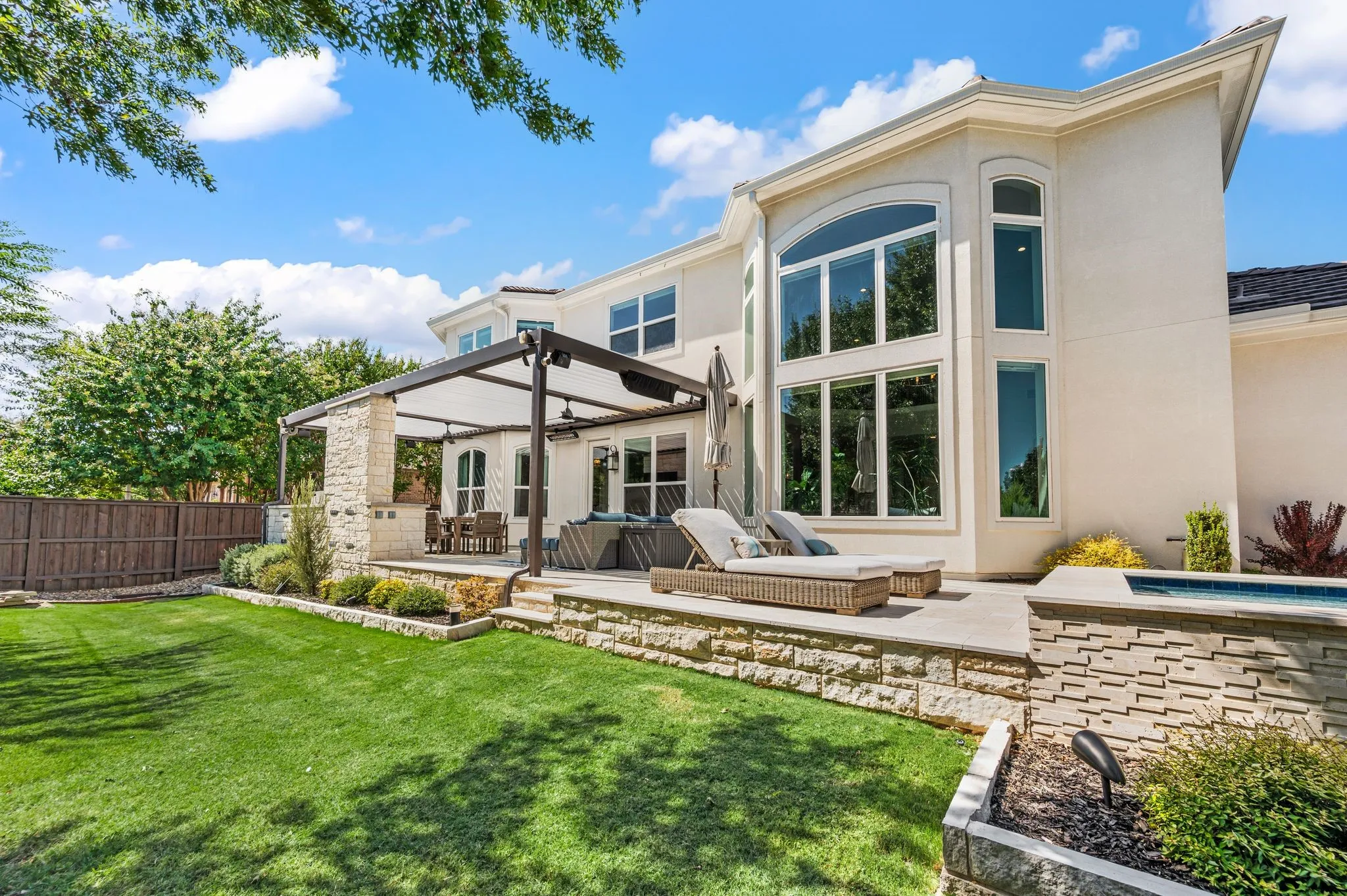 Modern residence featuring a striking two-story glass facade with expansive window arrays, stone and stucco exterior finishes, and a covered patio structure with integrated shade system