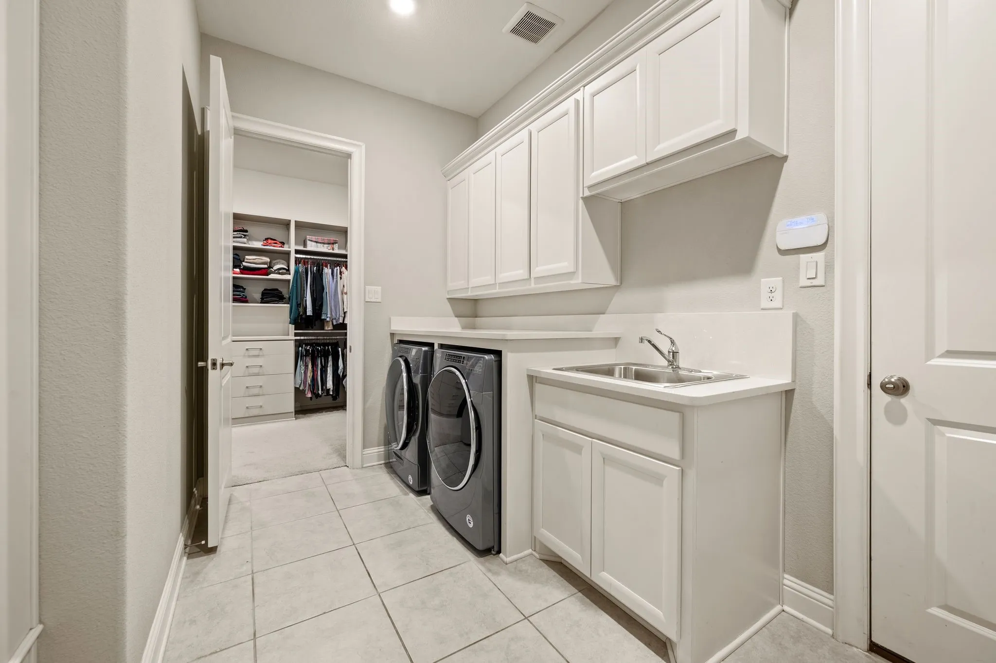 Dedicated laundry room featuring stacked front-load appliances, white shaker-style cabinetry with upper storage, tile flooring, and an integrated sink with countertop workspace