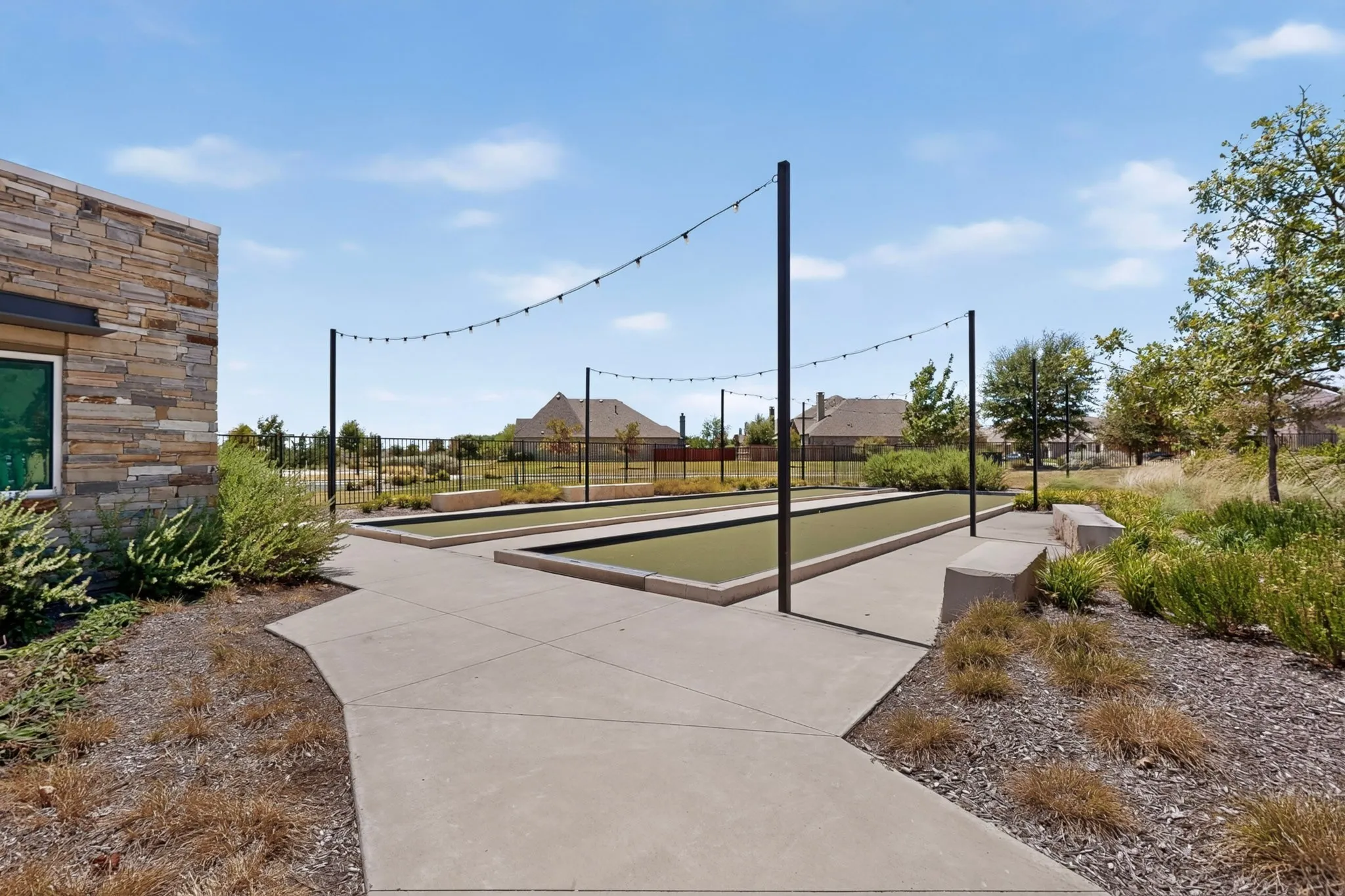 Two bocce ball courts, concrete pathways, stone-clad exterior building, permanent string lighting, and integrated bench seating