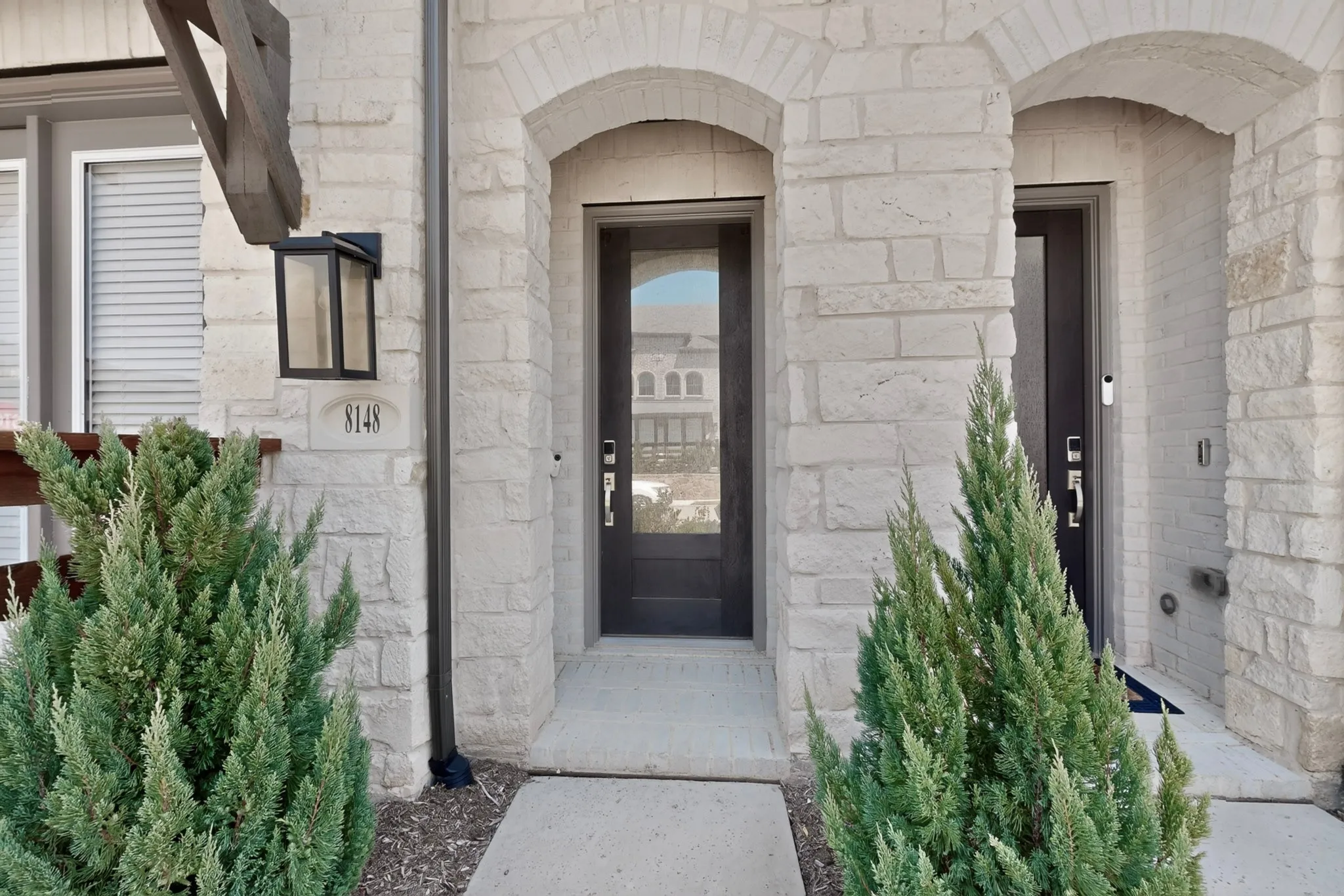 Stone facade with arched entryways, dark paneled entry door with glass insert, concrete walkway, and exterior wall lantern