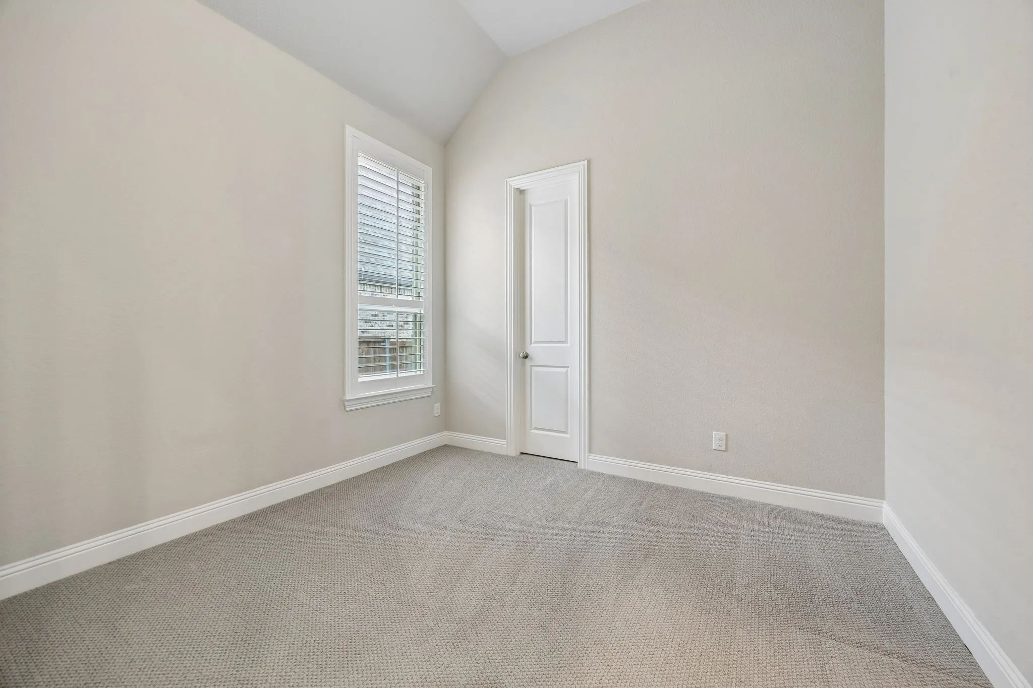 Vaulted ceiling, neutral wall paint, carpeted flooring, white trim, and a window with plantation shutters