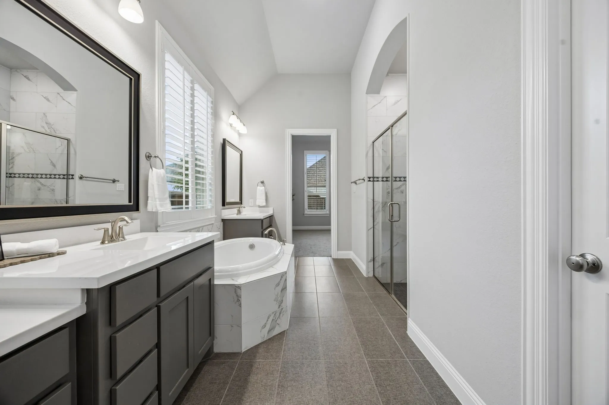 Dual vanity sinks with white countertops, corner soaking tub with tile surround, glass-enclosed shower with tiled walls, and large format tile flooring