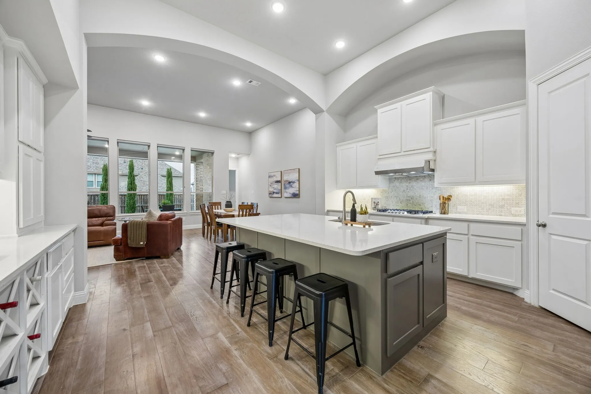 Open-concept kitchen with a large island, white shaker cabinetry, patterned tile backsplash, wood-finish flooring, and arched entryway