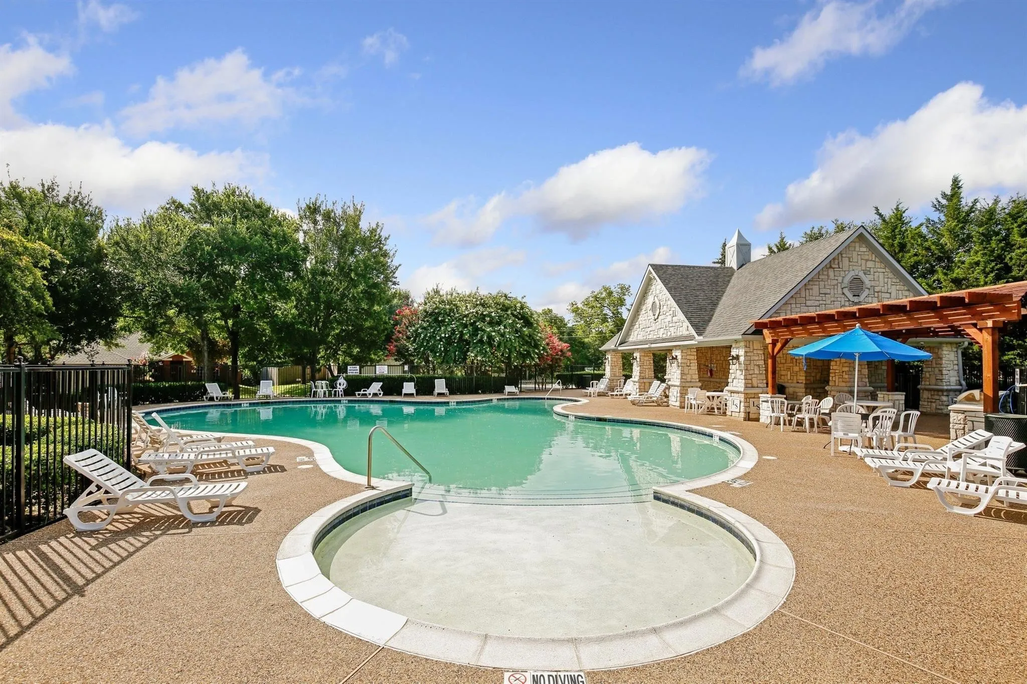 Large swimming pool with an integrated spa, surrounded by a textured concrete deck, a stone-clad clubhouse, and a wooden pergola structure