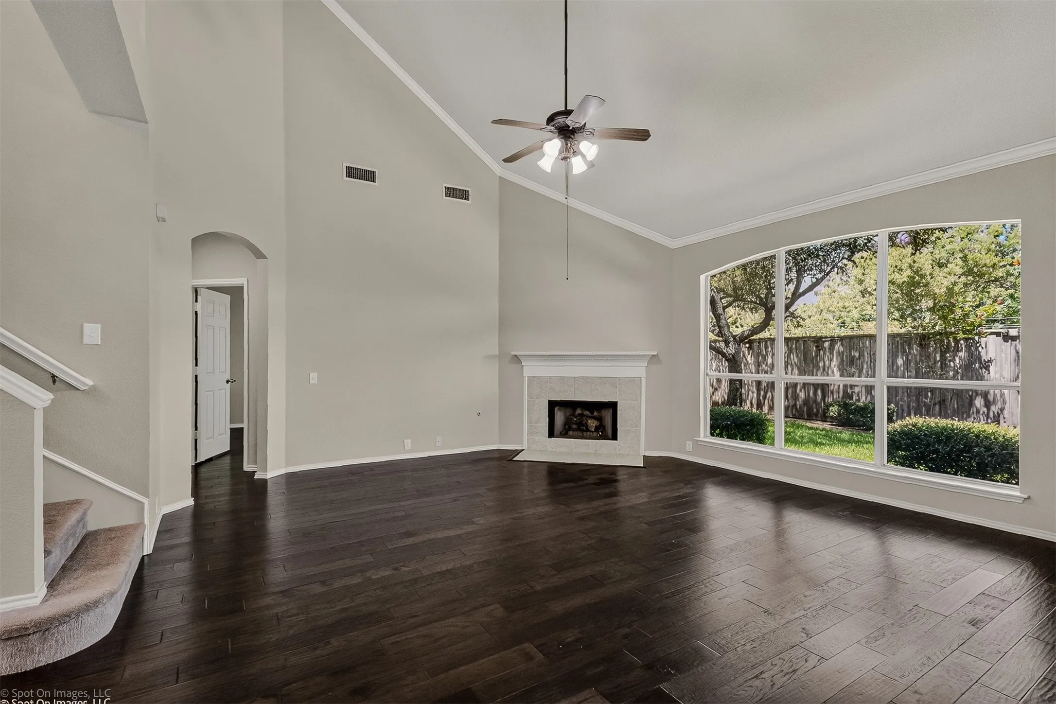 Vaulted ceiling with crown molding, dark wood-finish flooring, fireplace with stone surround, large bay window, and a ceiling fan with integrated lighting