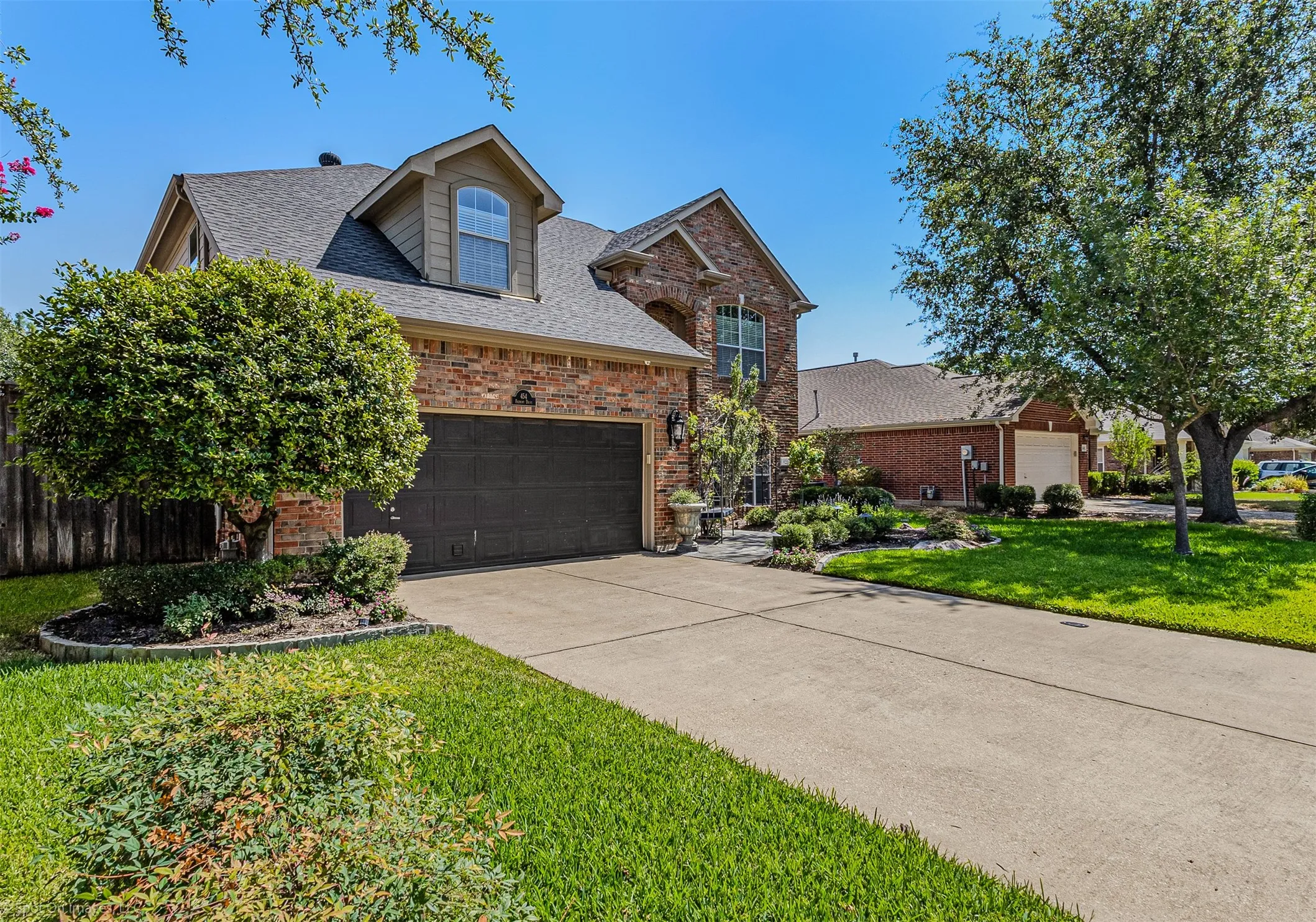 Brick exterior with dormer window, attached garage, concrete driveway, and established landscaping