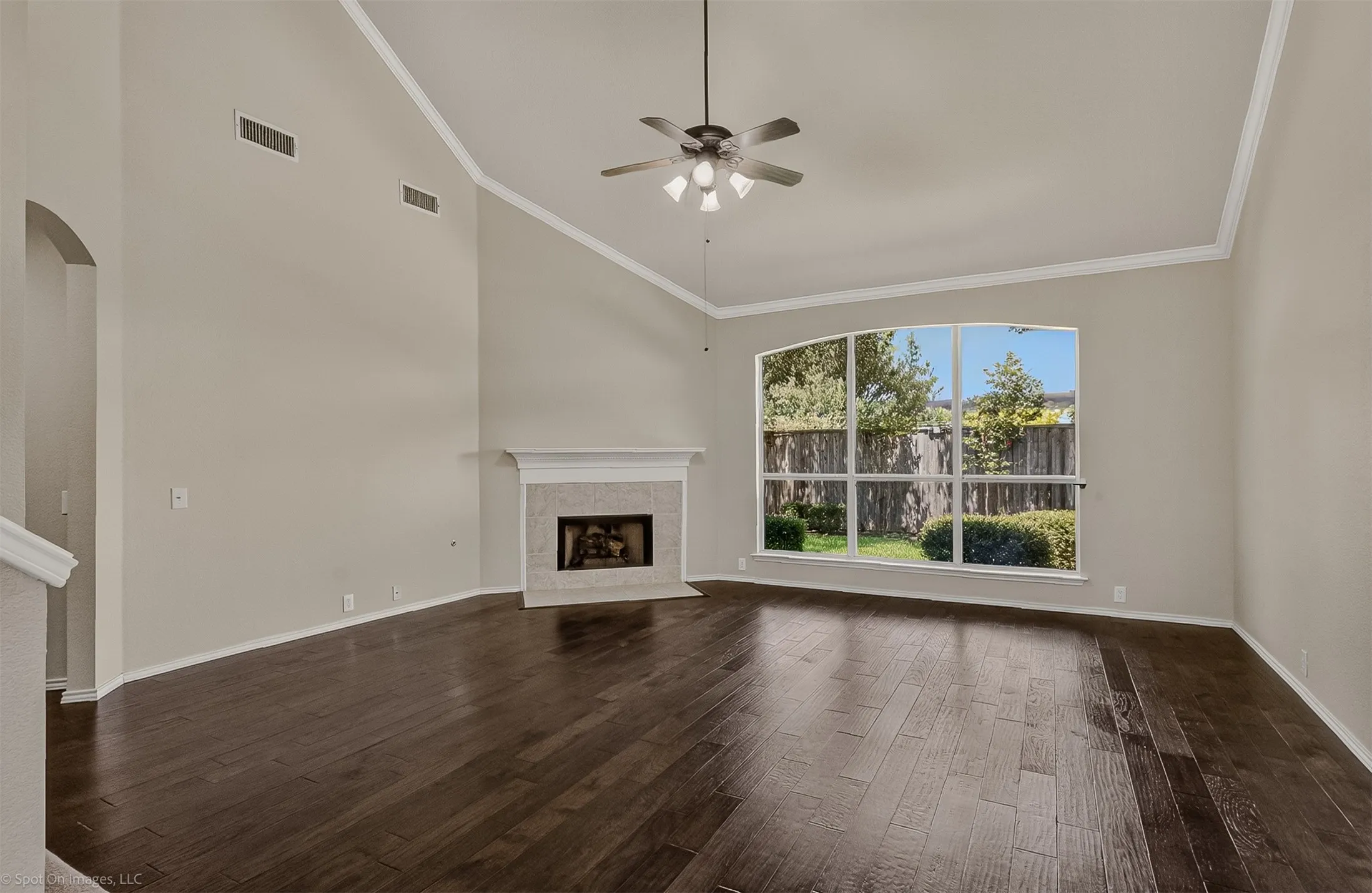Vaulted ceiling with crown molding, dark wood-finish flooring, fireplace with a tile surround, large arched window, and a ceiling fan with integrated lighting