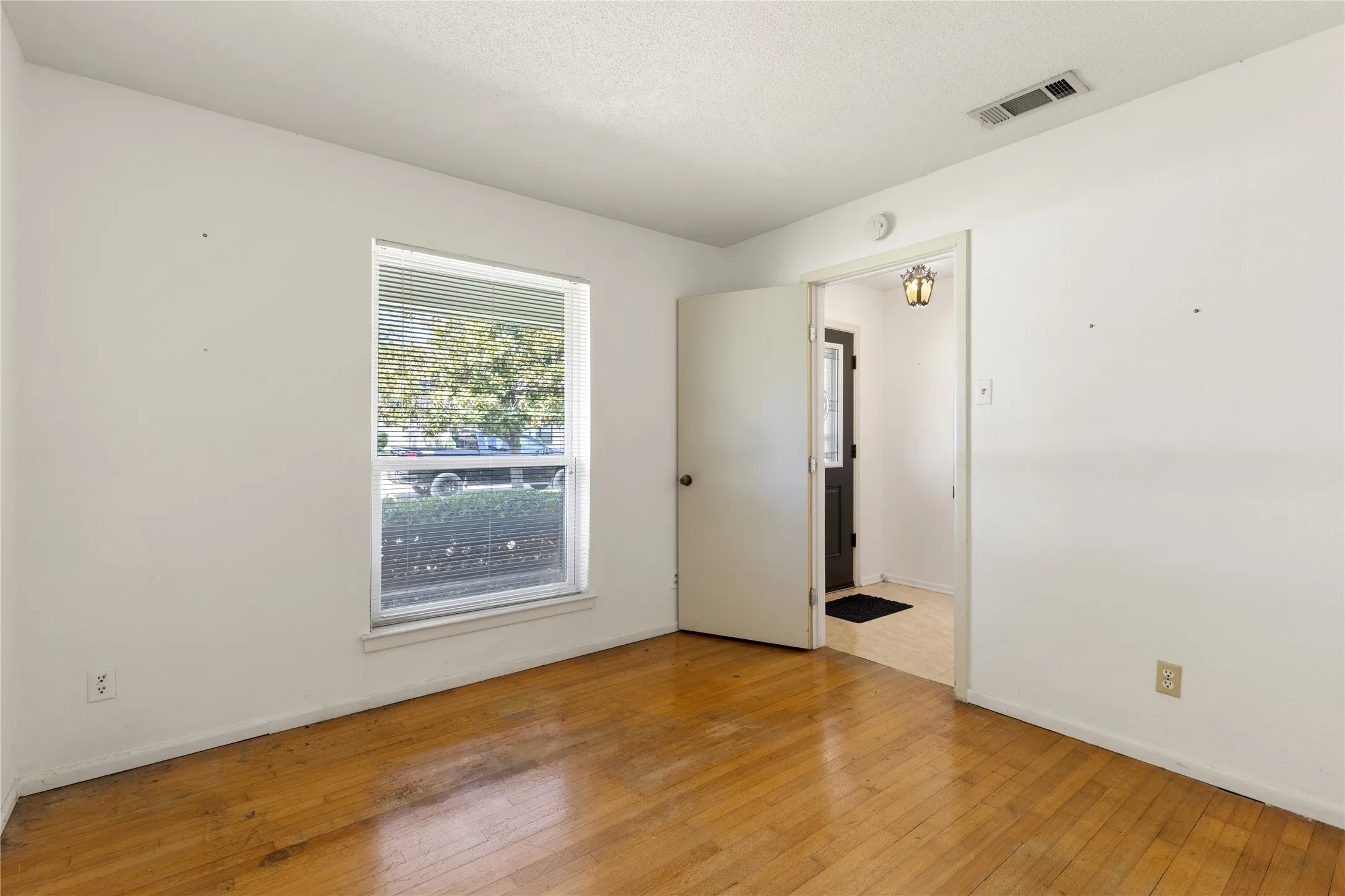 Hardwood flooring, white painted walls, single window with blinds, overhead lighting, and an open doorway