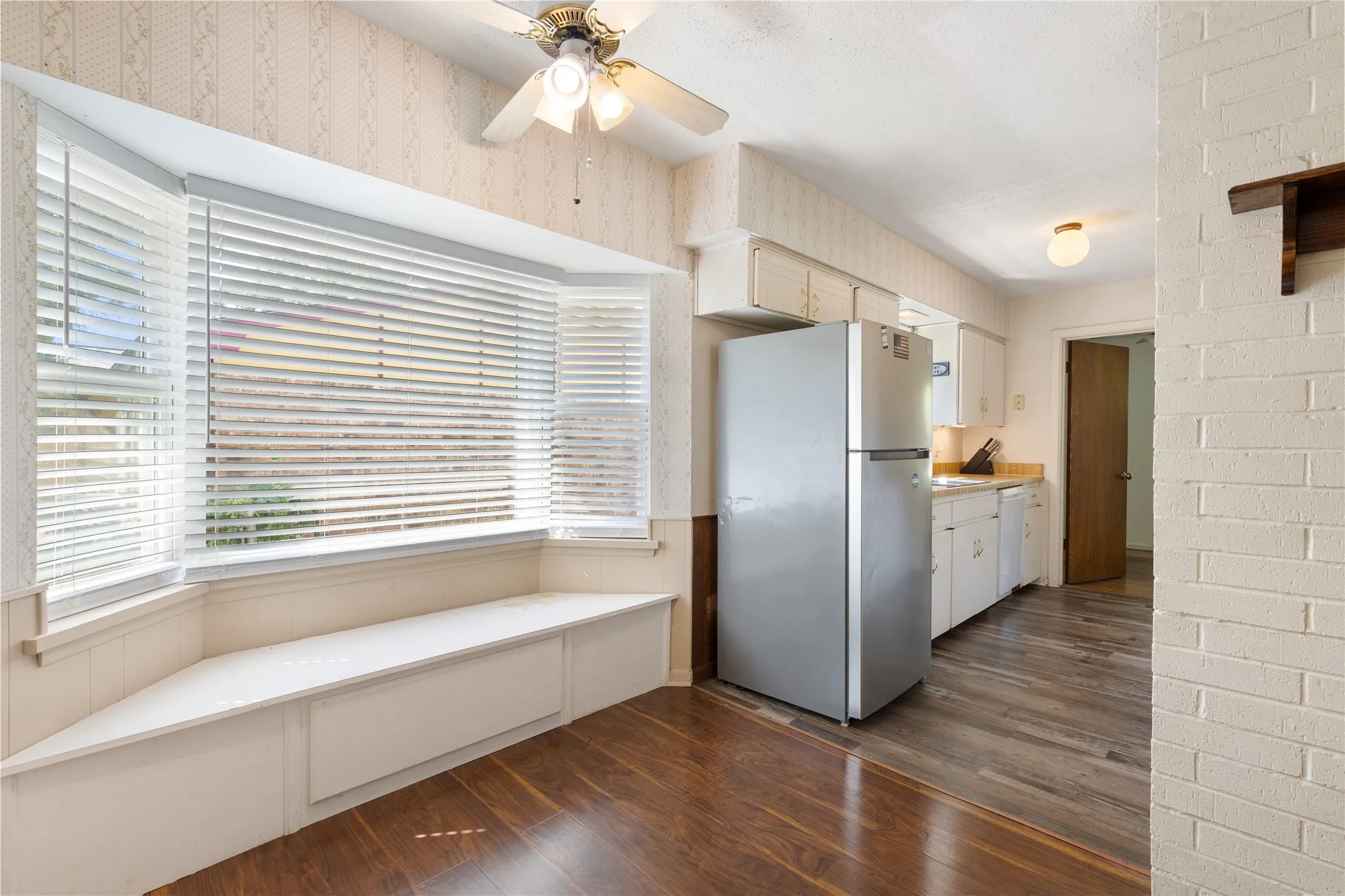 Bay window with integrated bench seating, wood-finish flooring, white brick wall accent, ceiling fan, and white cabinetry