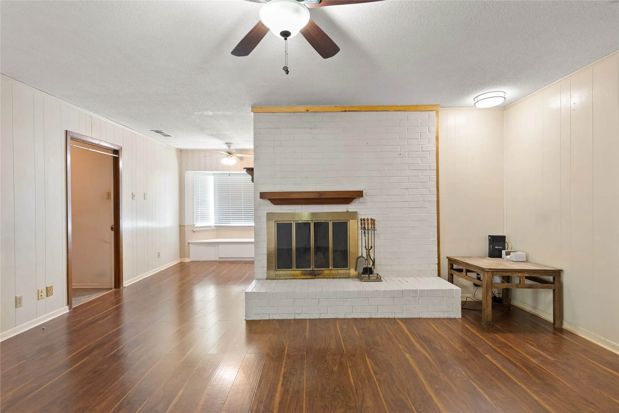 Expansive room featuring wood-finish flooring, a white brick fireplace with a wood mantle, and a ceiling fan with light