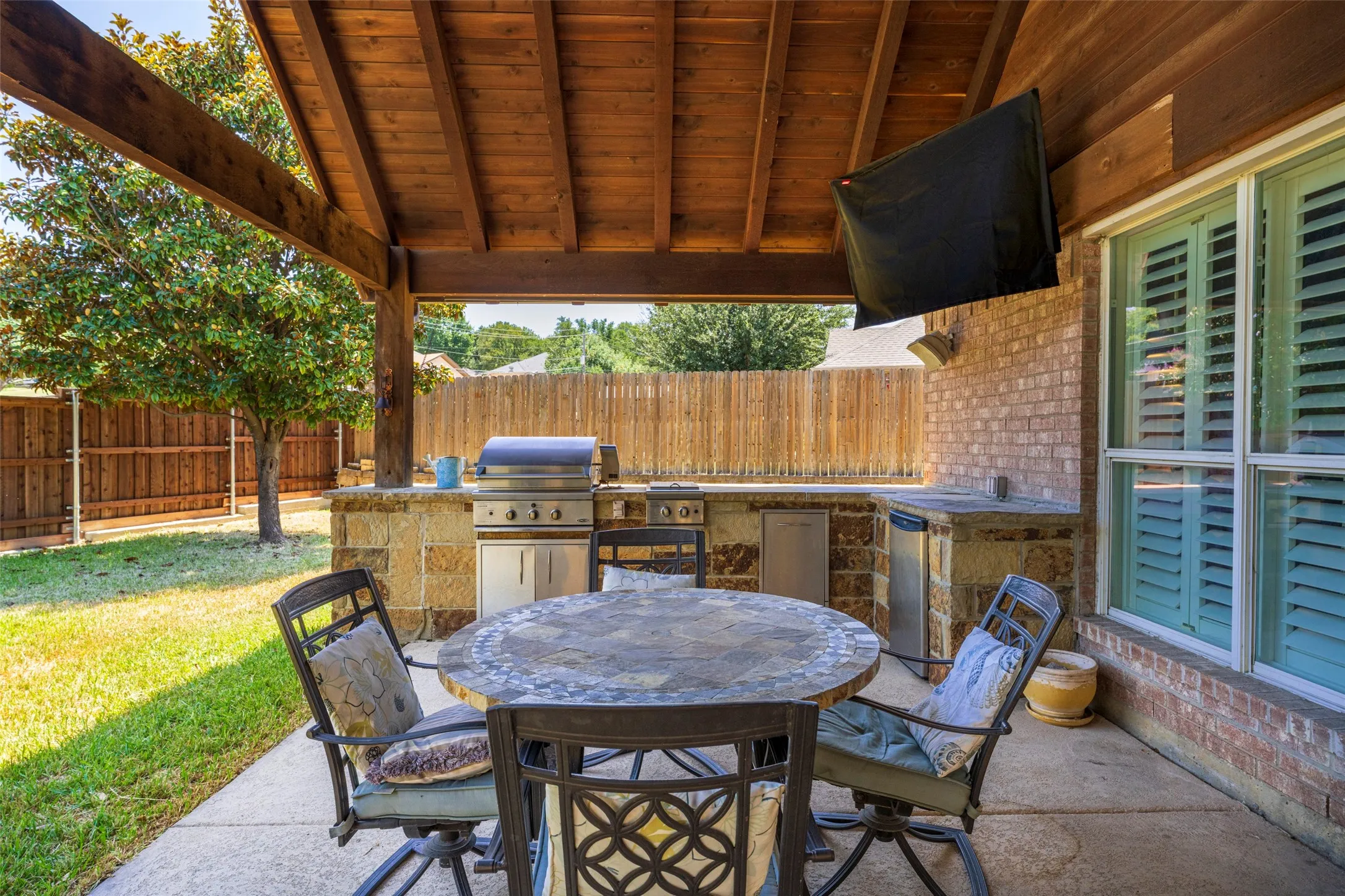 Covered patio featuring a built-in outdoor kitchen with grill, brick exterior, wood-plank ceiling, and privacy fencing
