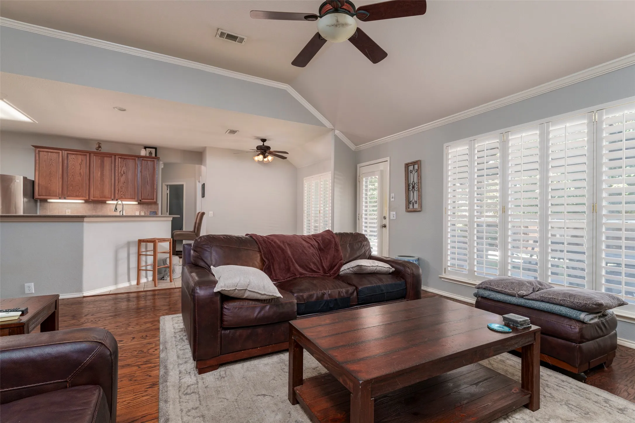 Vaulted ceiling with a ceiling fan, wood-finish flooring, kitchen pass-through with wood cabinetry, and large windows with plantation shutters
