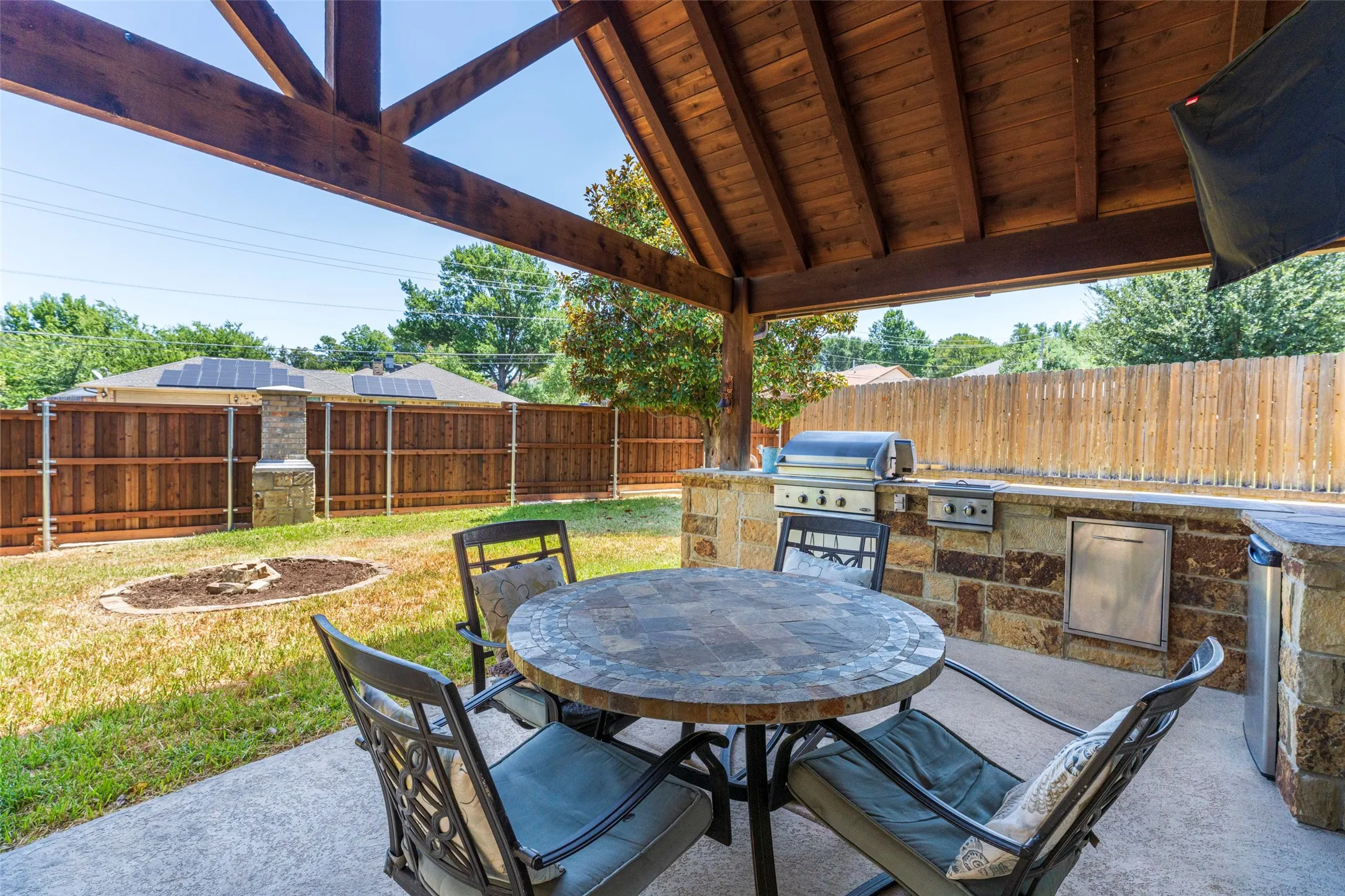 Covered patio with exposed wood beam ceiling, built-in stone outdoor kitchen with grill, and concrete flooring