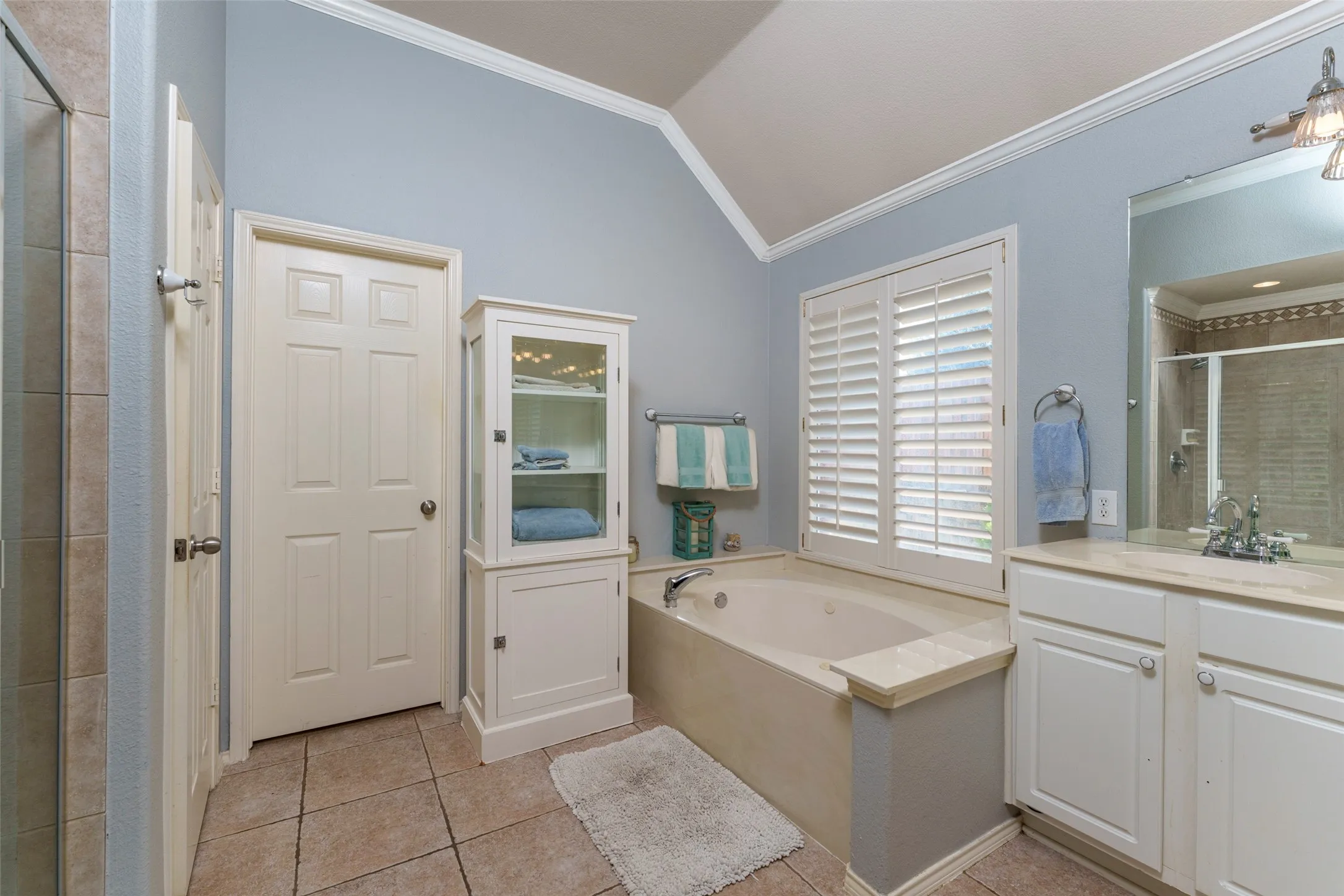 Vaulted ceiling, tiled flooring, built-in soaking tub, window with plantation shutters, and a single vanity with a mirror