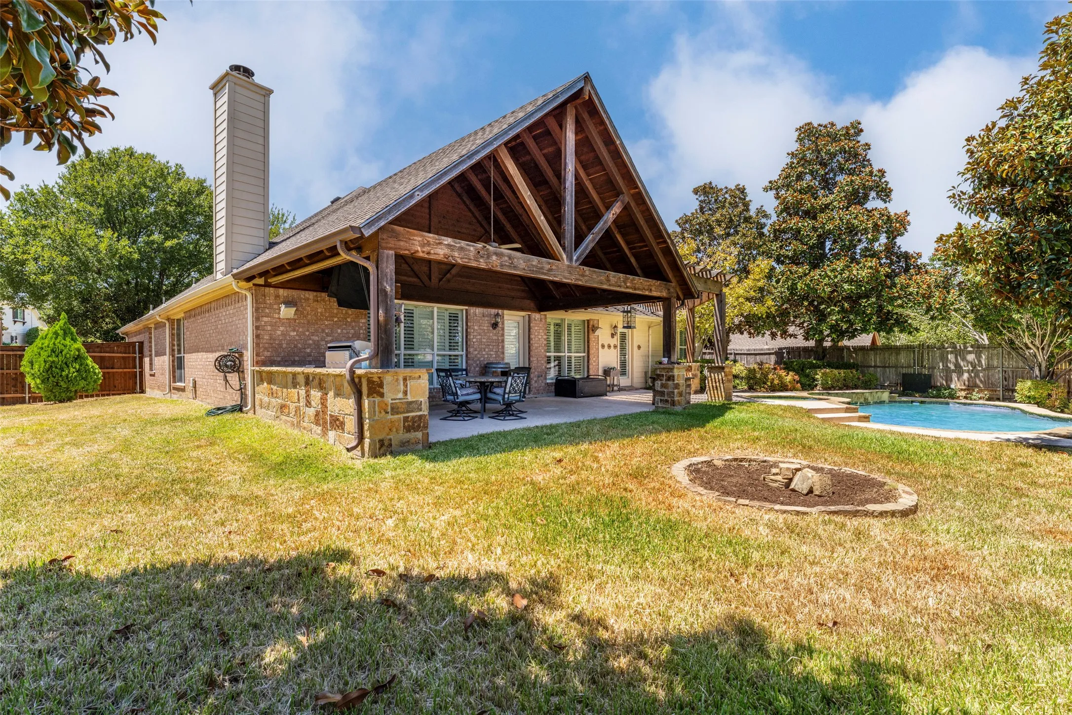 Expansive backyard featuring a covered patio with exposed wood beams, a built-in swimming pool, a brick exterior, and a dedicated fire pit area