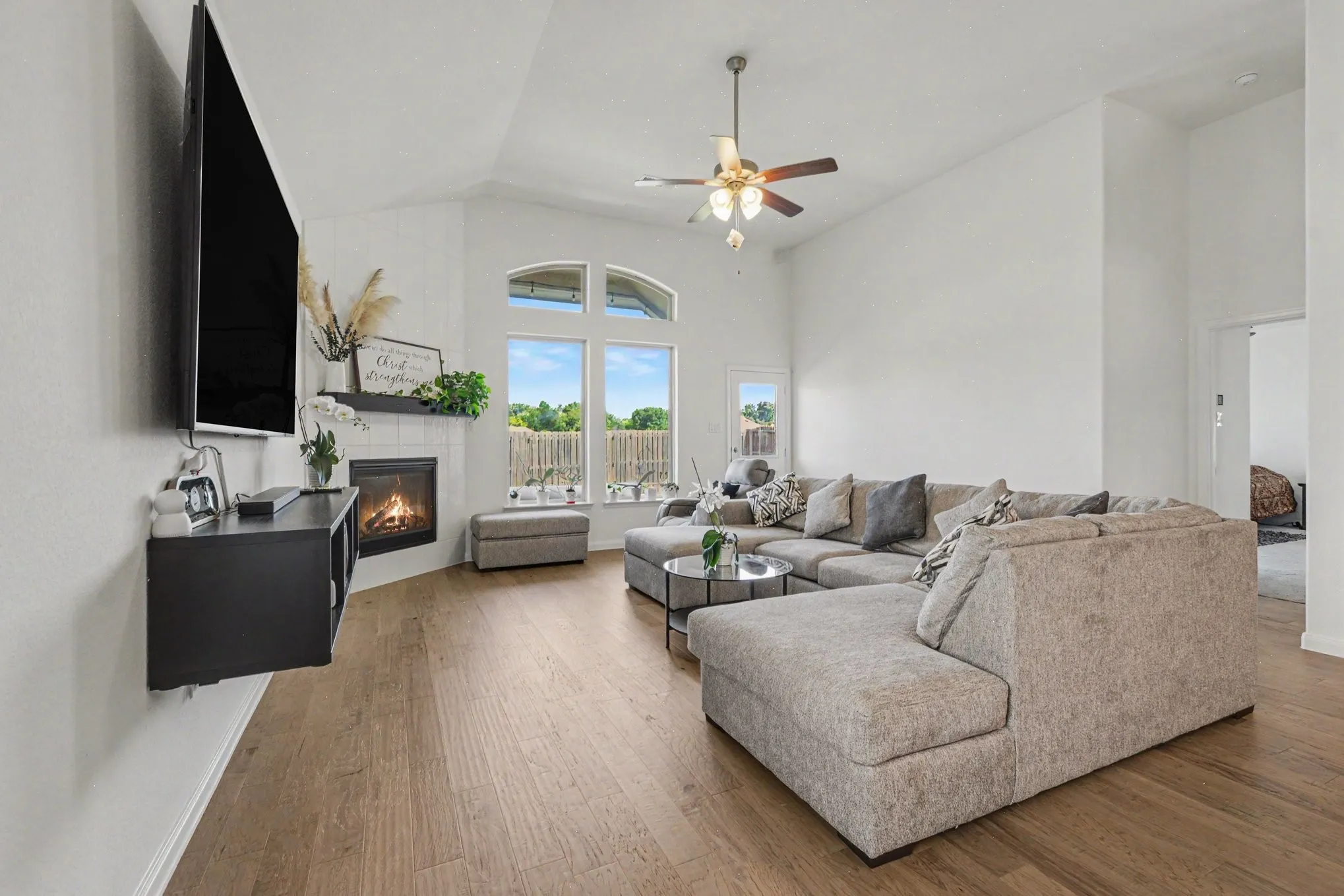 Vaulted ceiling with ceiling fan, wood-finish flooring, corner fireplace with white tile surround, and arched windows