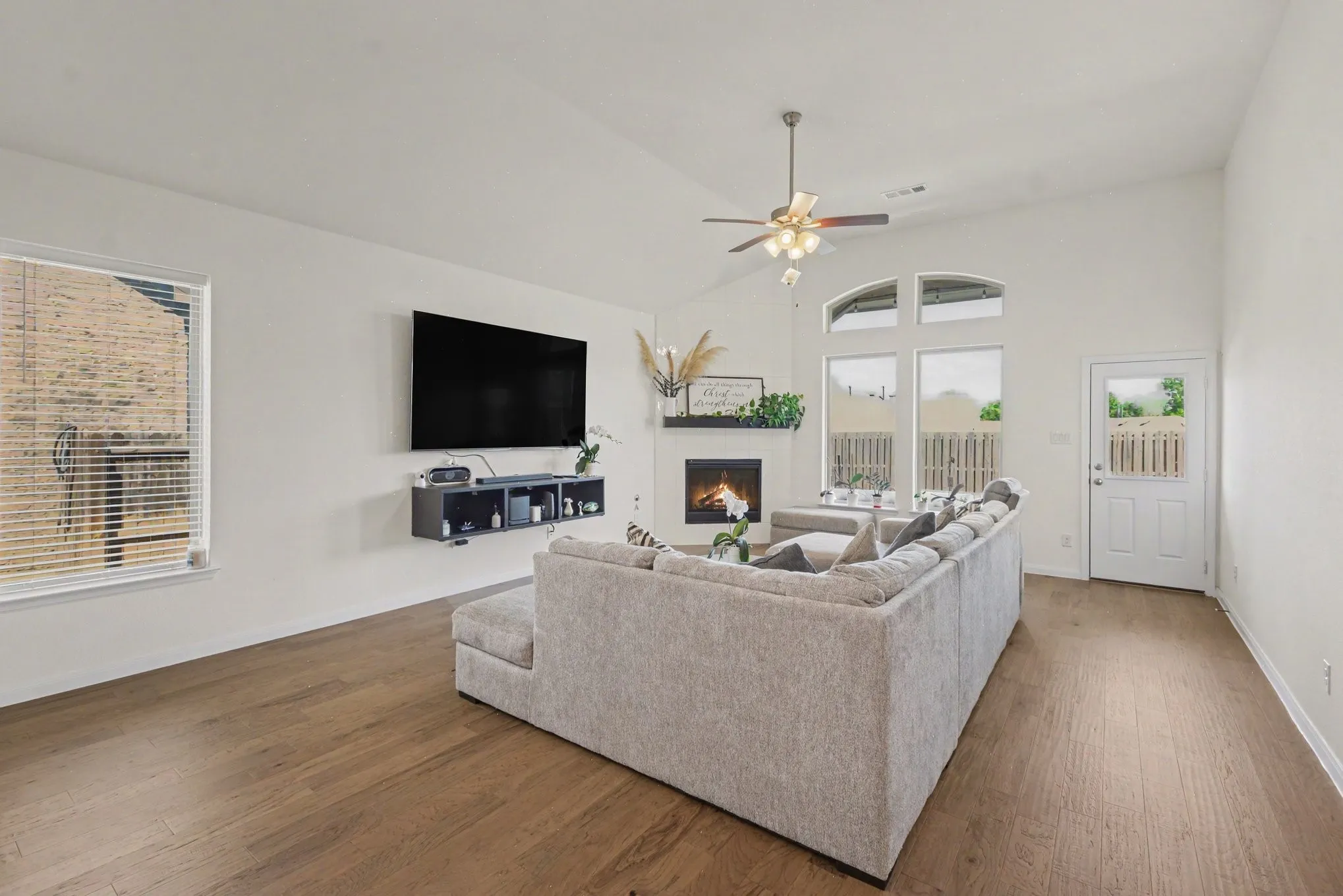 Vaulted ceiling, wood-finish flooring, corner fireplace with mantel, ceiling fan with light fixture, and arched window