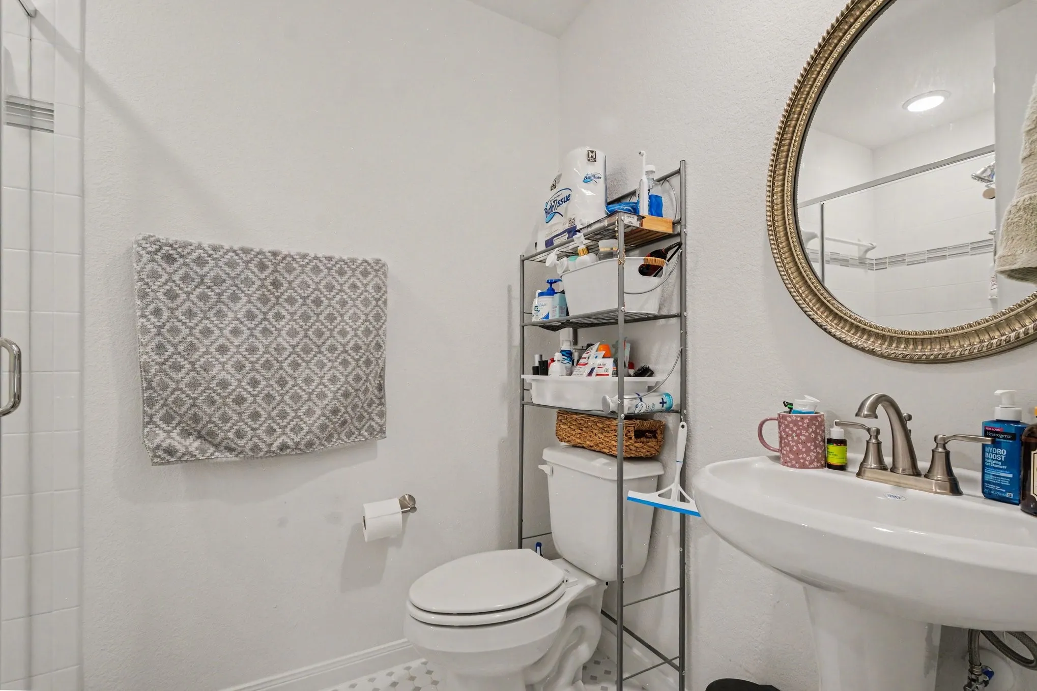 Pedestal sink with a brushed nickel faucet, a toilet, a decorative round mirror, and a shower with a clear glass enclosure