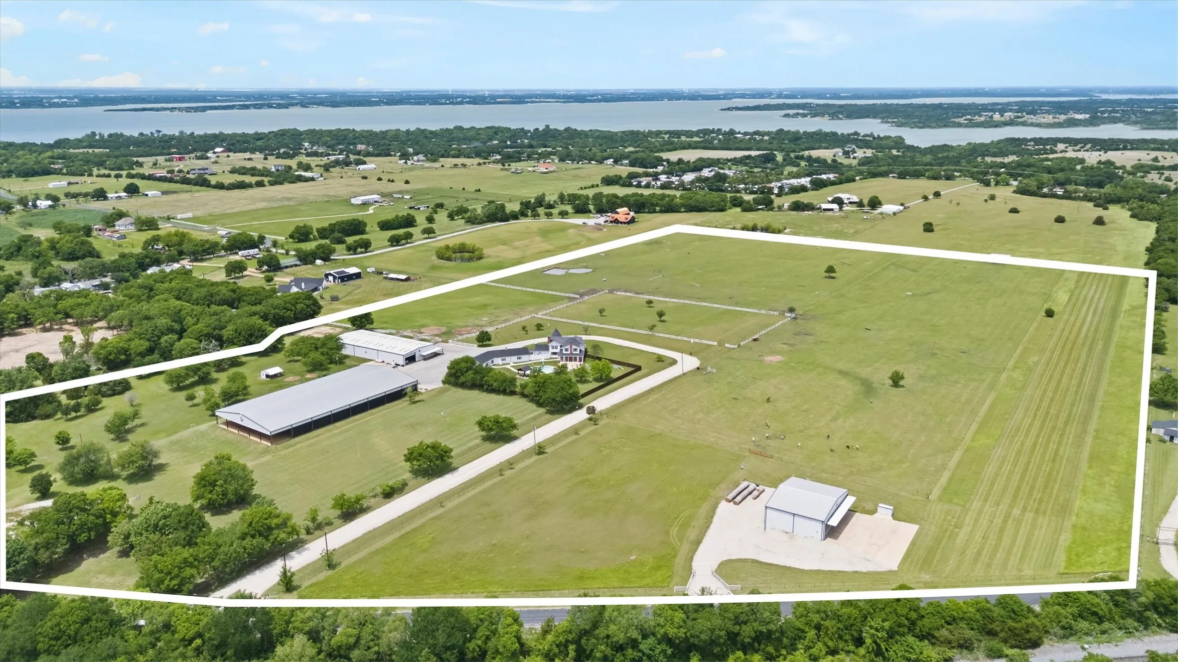 Expansive rural property featuring a large body of water in the background, a substantial primary residence, an extensive covered arena, multiple outbuildings, and open fields with scattered trees
