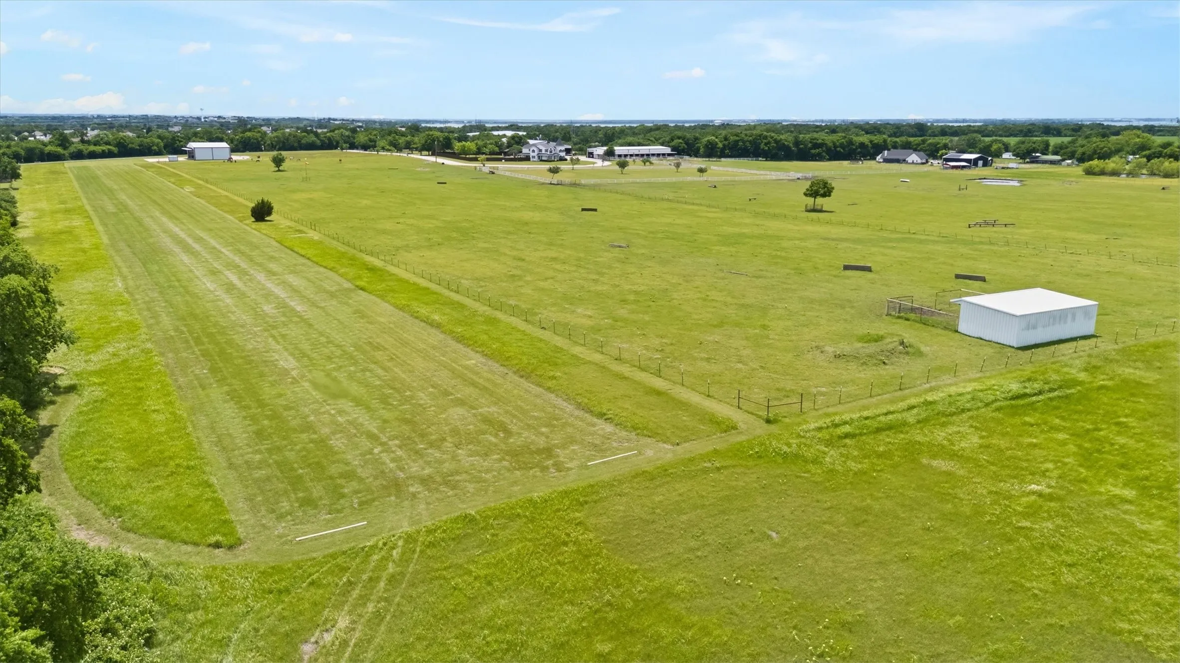 Expansive grass field featuring a mowed strip, bordered by scattered trees and fencing