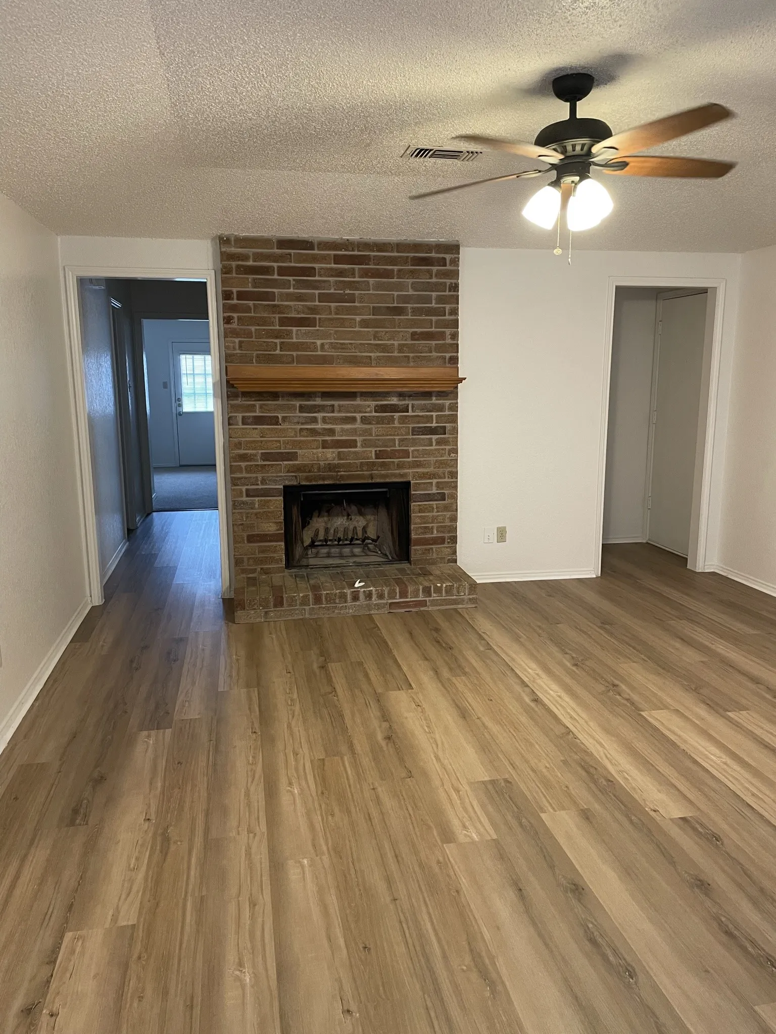 Wood-finish flooring throughout, brick fireplace with a wood mantel, and a ceiling fan with integrated lighting