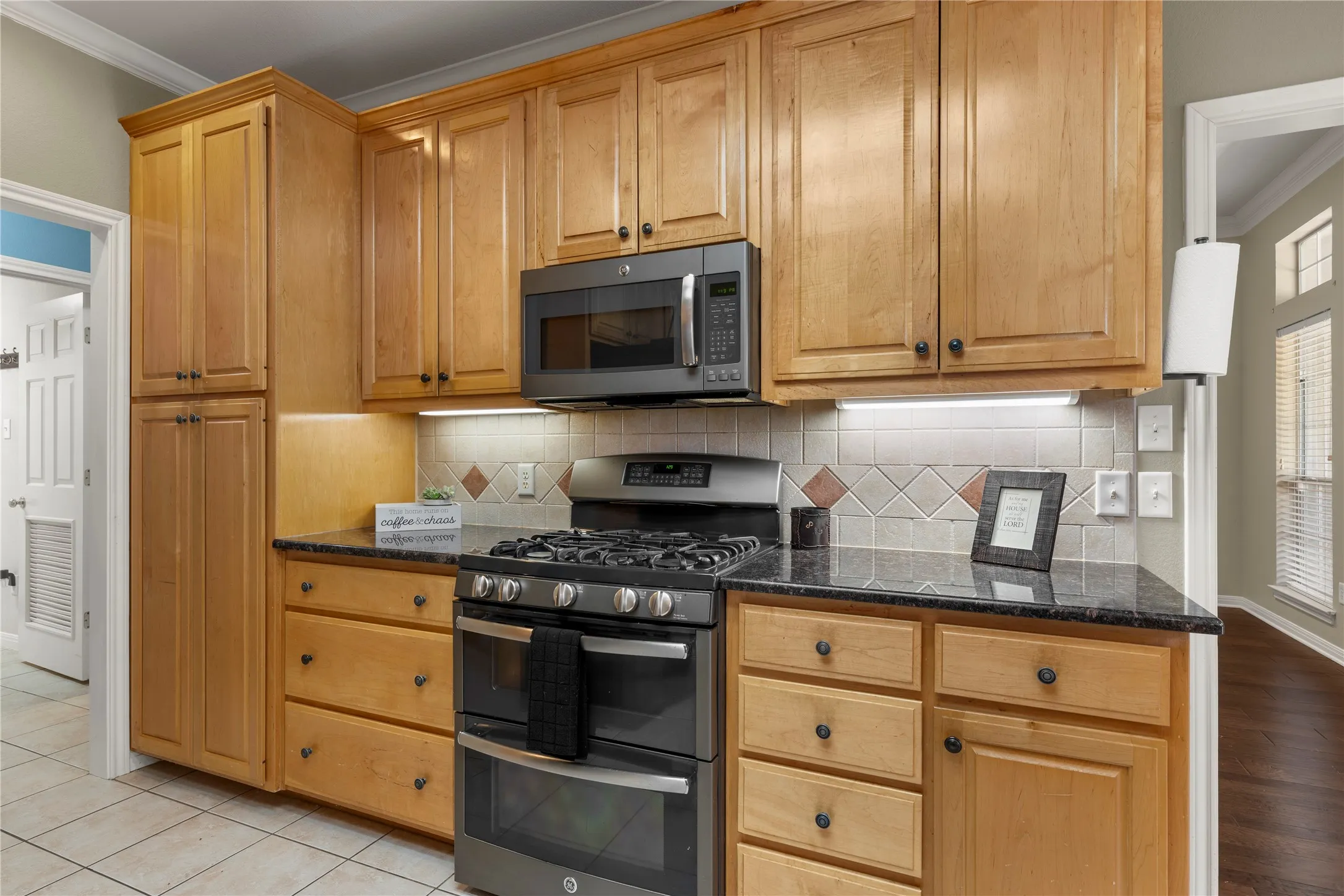 Warm wood cabinetry with abundant storage and brushed hardware anchors this kitchen, which features stainless steel dual-fuel range with six burners, matching microwave, and black granite countertops