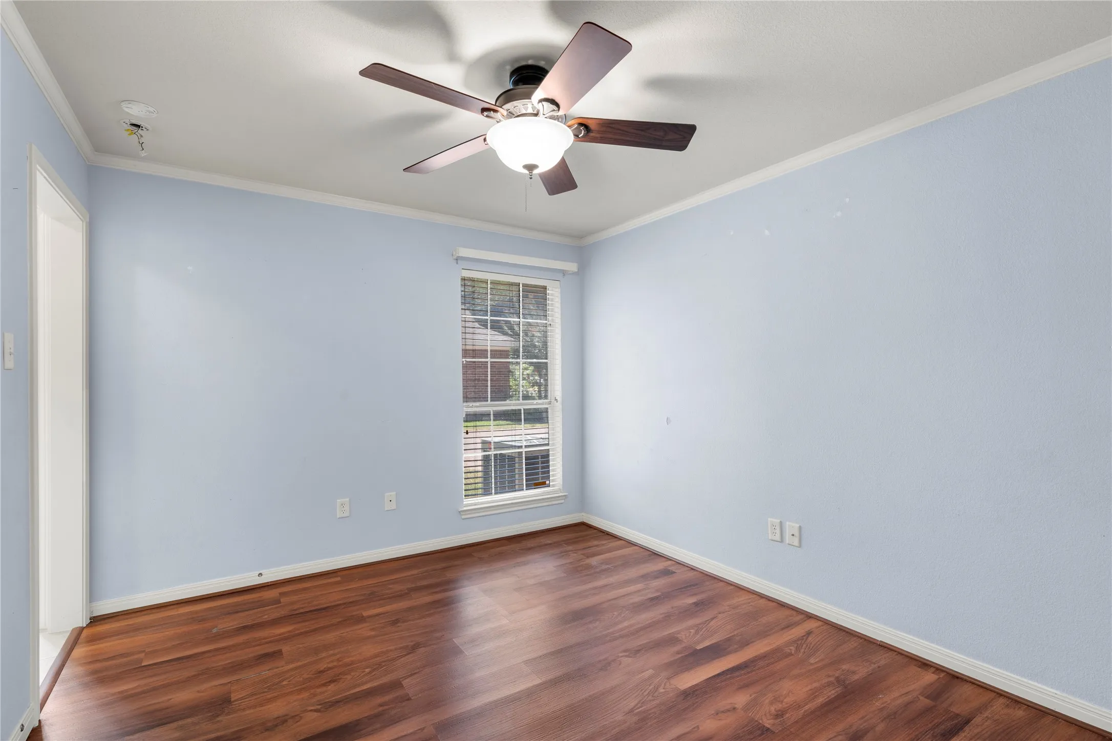 Wood-finish flooring, single window with blinds, ceiling fan with integrated lighting, and white trim accents
