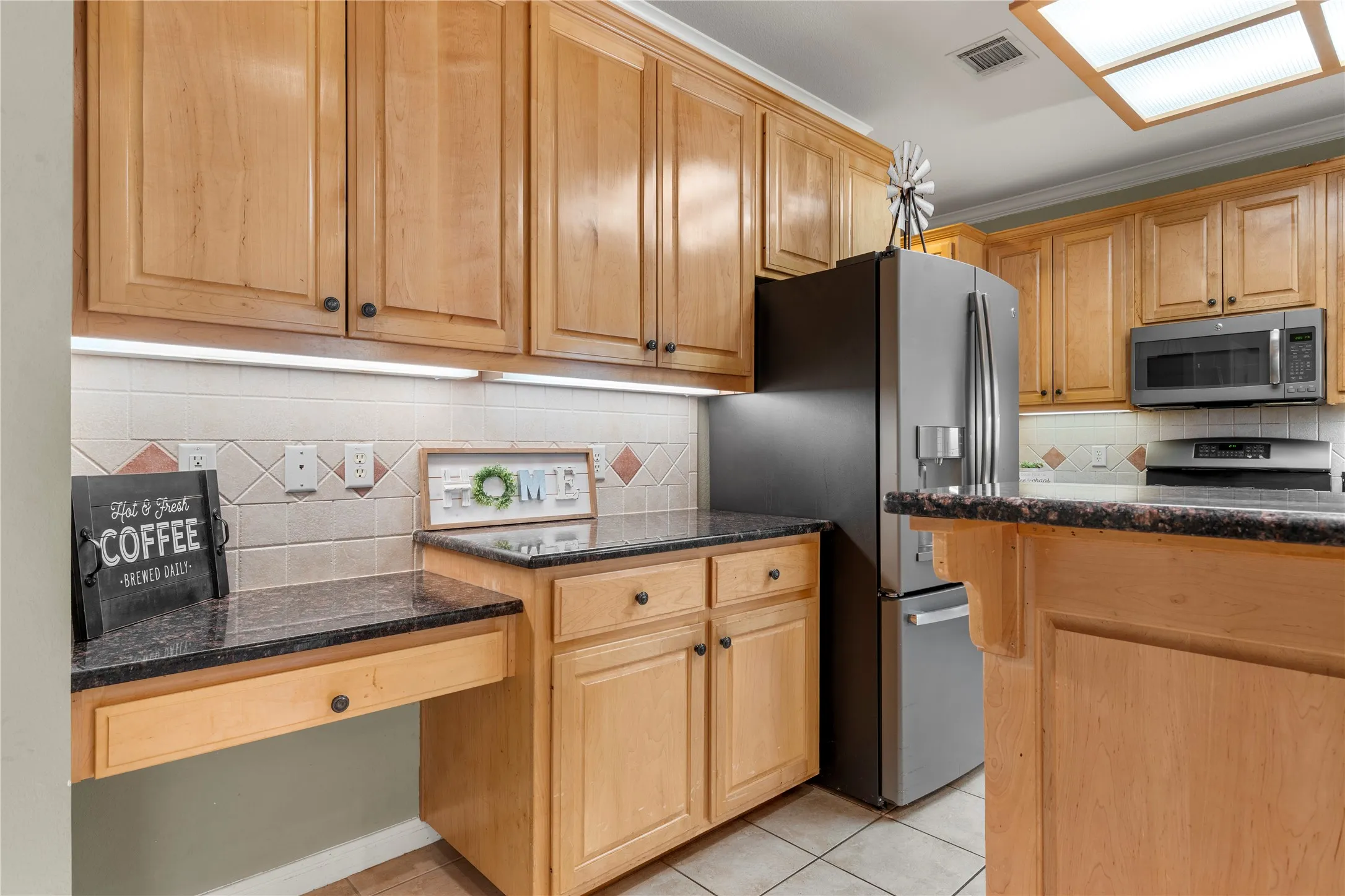 Wood-finish cabinetry, dark speckled countertops, tile flooring, stainless steel appliances, and a built-in desk area