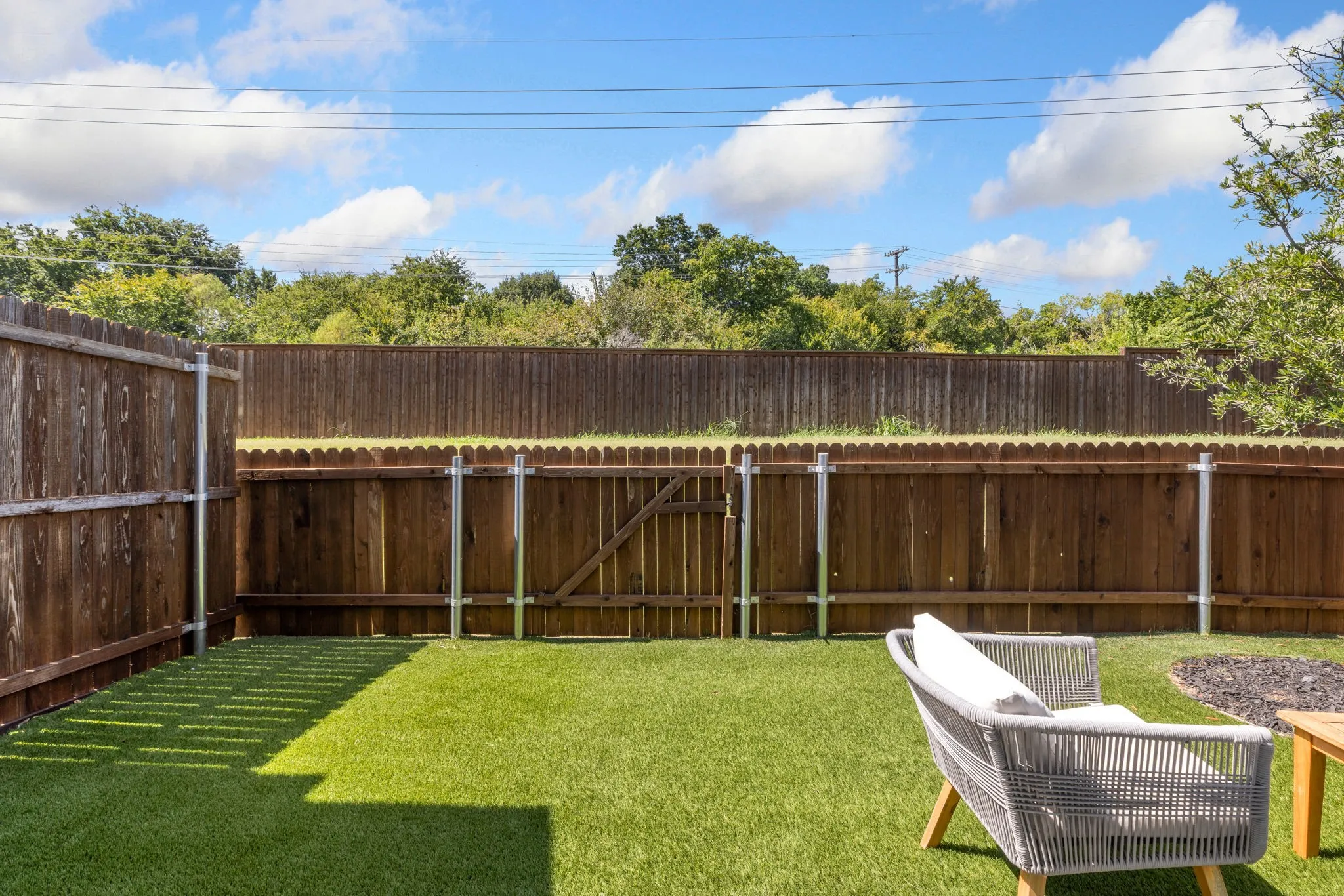Expansive backyard featuring manicured artificial turf, wood-finish fencing with covered structure, mature tree backdrop, and modern outdoor seating area with yellow accent elements