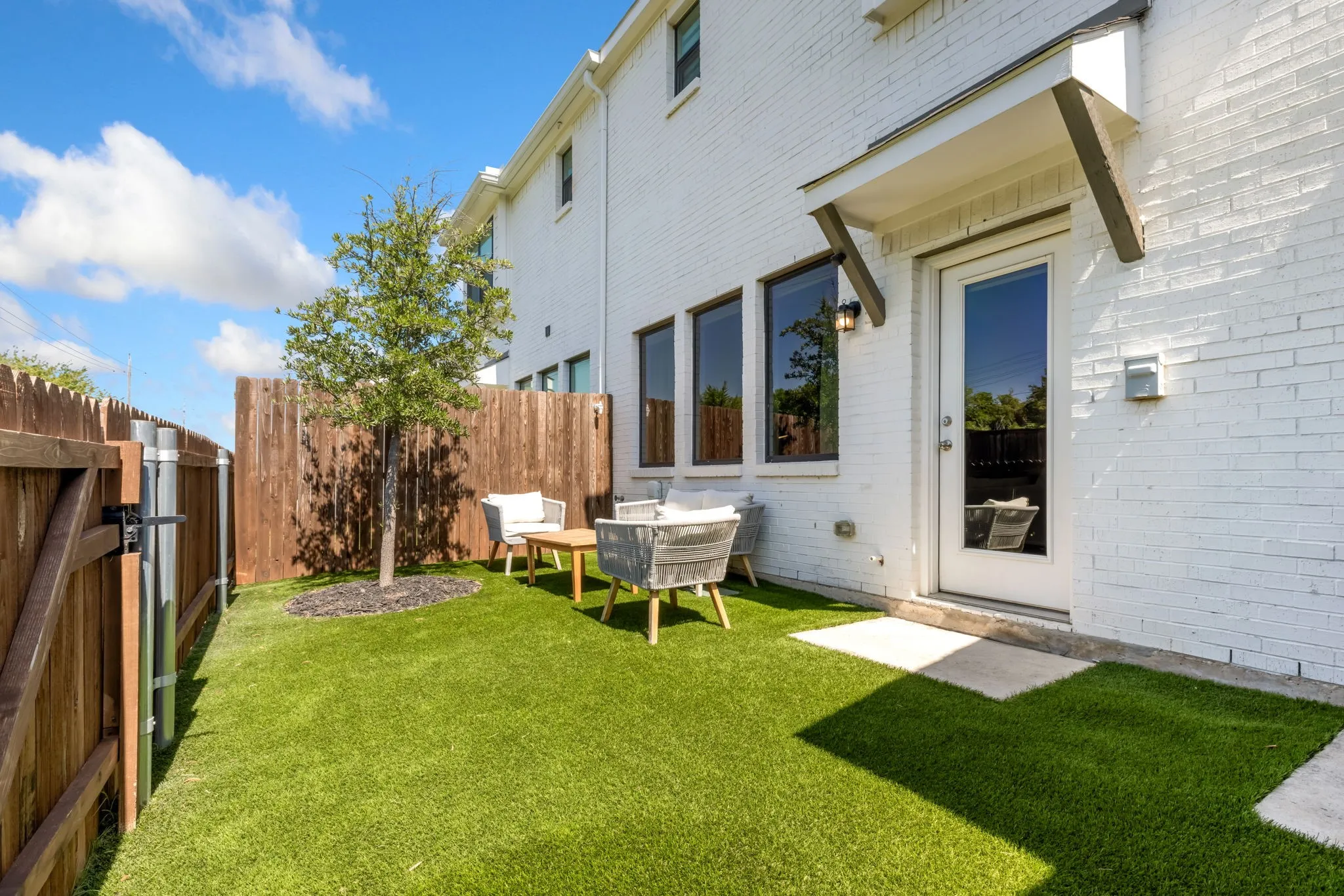 Bright, light-filled backyard with manicured lawn and mature landscaping, anchored by a modern white brick residence featuring multi-pane glass doors, black-trimmed windows, and covered entry