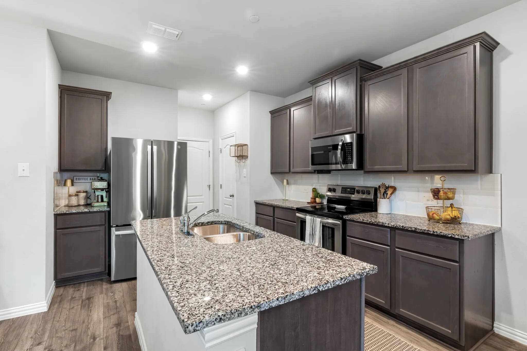 Modern kitchen featuring dark wood cabinetry, speckled quartz countertops, a large center island with seating space, stainless steel appliances, recessed ceiling lighting, and wood-finish flooring throughout the open layout