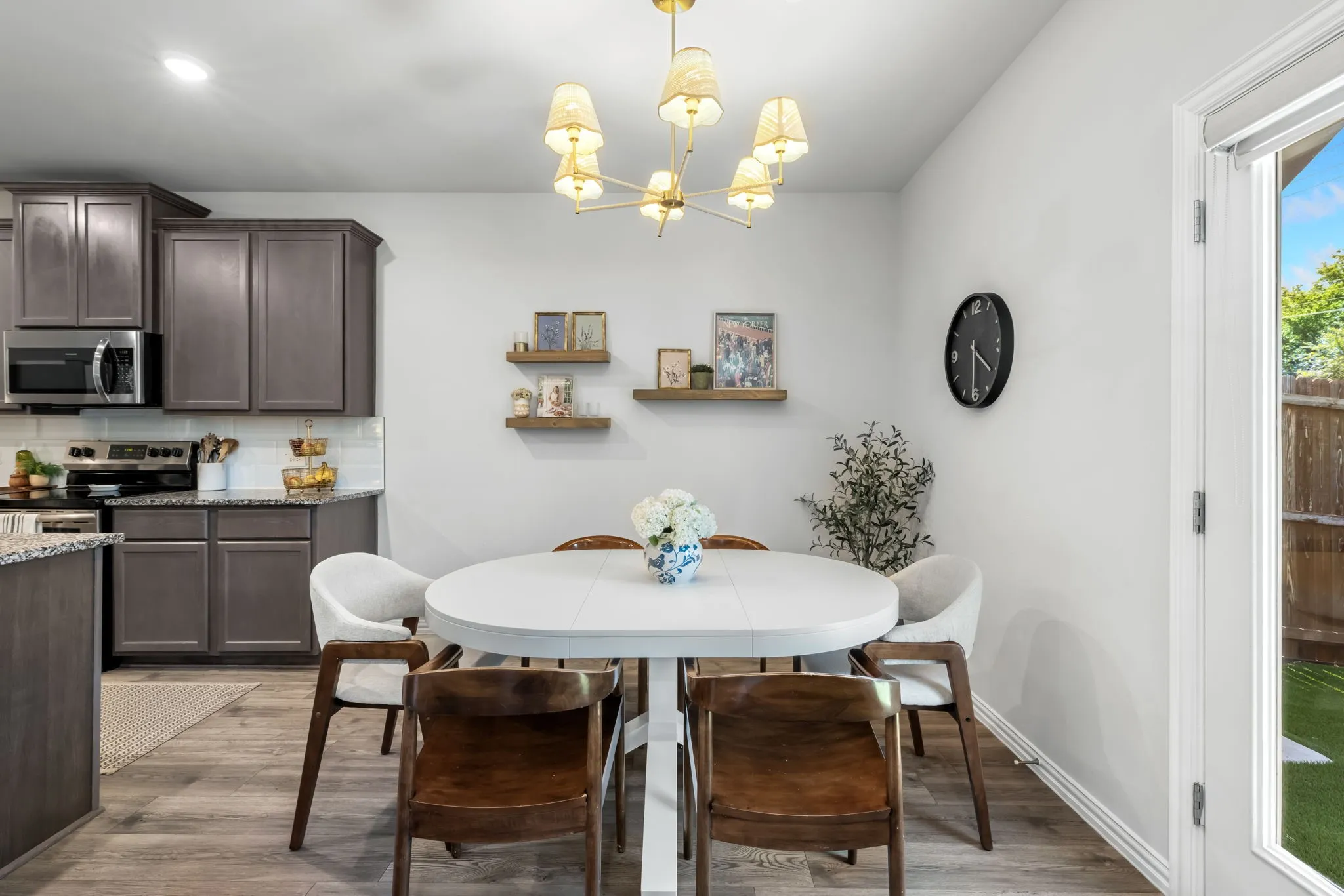Modern kitchen and dining area featuring stainless steel appliances, dark cabinetry with ample storage, wood-finish flooring throughout, a round white dining table with seating for four, and a brass multi-pendant light fixture suspended above