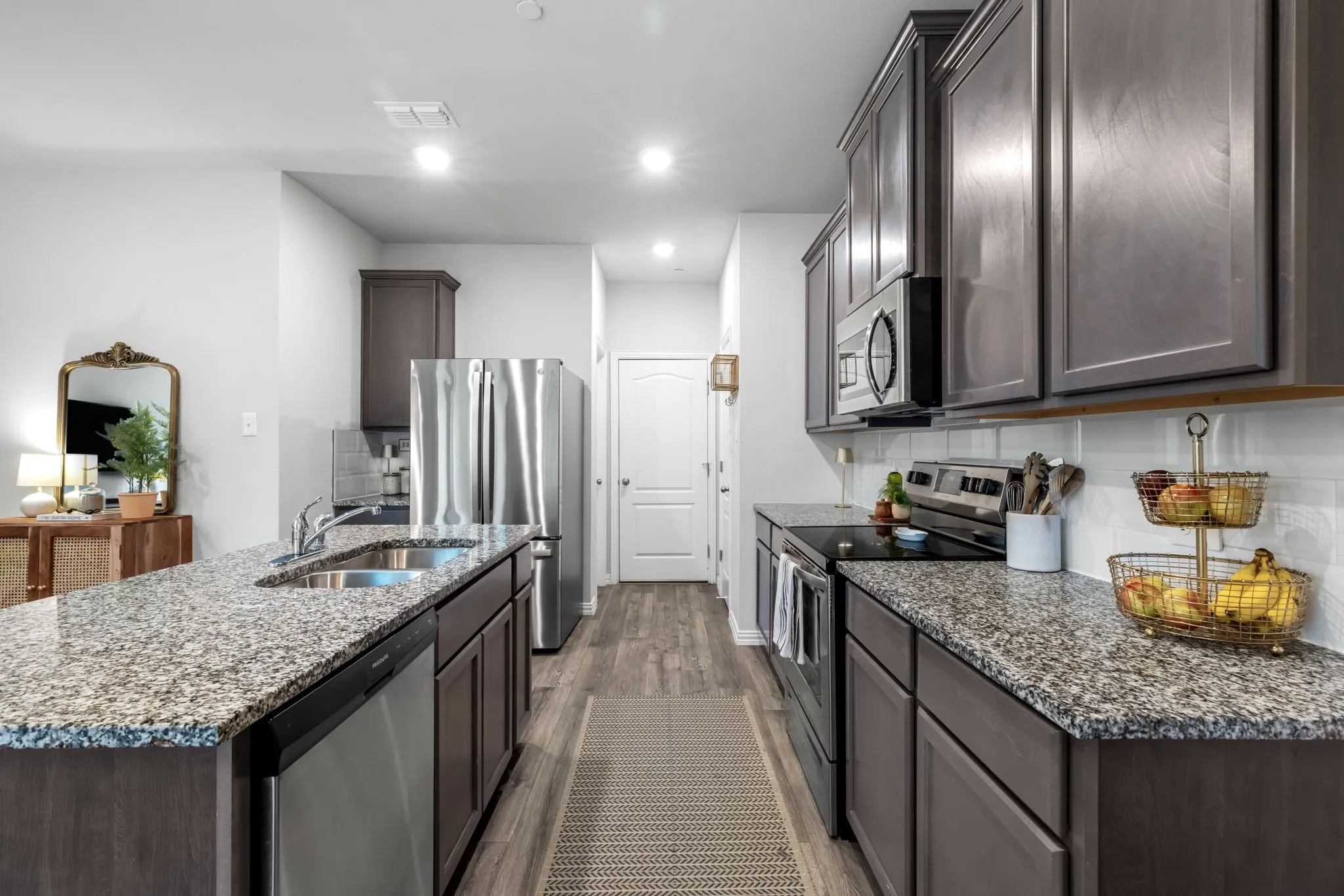 Galley-style kitchen featuring dark cabinetry with brushed metal hardware, speckled stone countertops, stainless steel appliances including a refrigerator and range, recessed ceiling lighting, and wood-finish flooring throughout