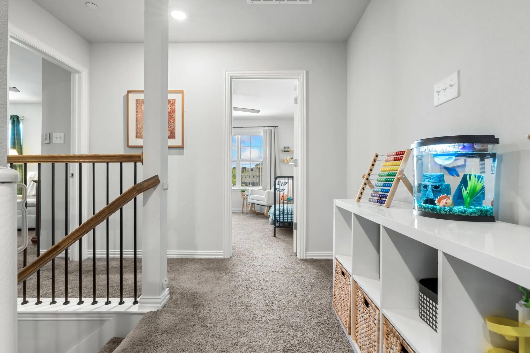 Bright upper-level hallway featuring neutral wall finishes, gray carpeting, and a wood-finish stair rail with metal balusters