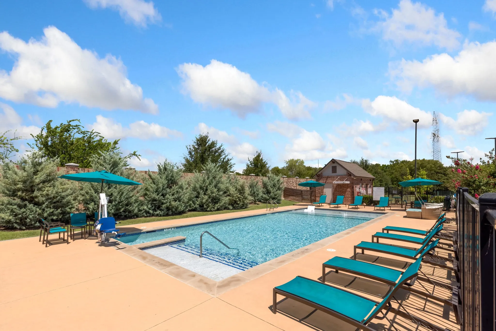 Resort-style recreational area featuring a sparkling saltwater pool with integrated shallow entry, expansive concrete decking with lounge seating, a covered pavilion with turquoise accents, and mature landscaping with evergreen trees providing natural screening