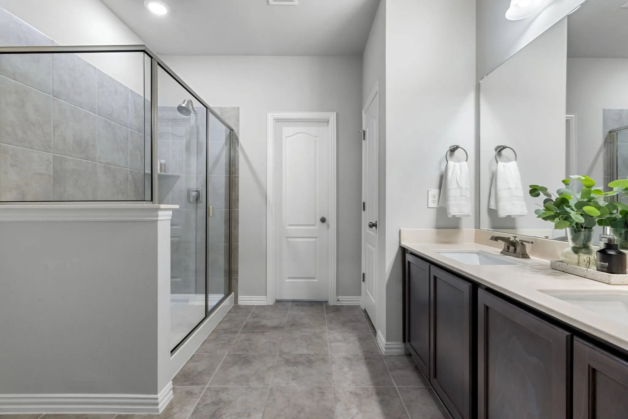 Spa-inspired primary bathroom featuring a spacious walk-in shower with frameless glass enclosure and tile surround, dual-sink vanity with dark cabinetry and quartz countertop, large framed mirror with integrated lighting, and porcelain tile flooring throughout