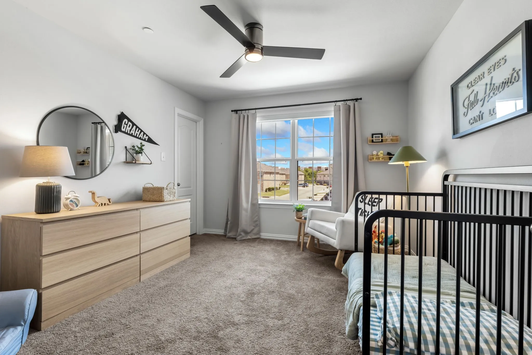 Bright nursery featuring expansive windows with natural light, light wood-finish dresser with round mirror and task lighting, black metal crib, ceiling fan, neutral carpet flooring, and scenic outdoor views