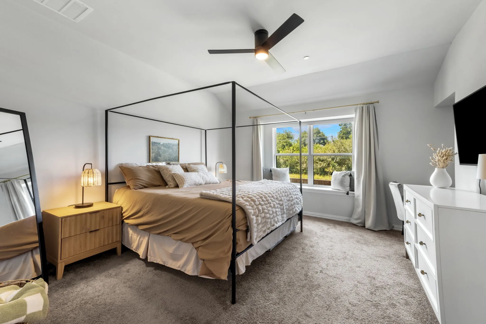 Spacious primary bedroom featuring a black metal four-poster bed frame, wood-finish nightstands with warm lighting, white built-in cabinetry, ceiling fan with integrated lighting, carpet flooring, and large windows with natural light and outdoor scenery
