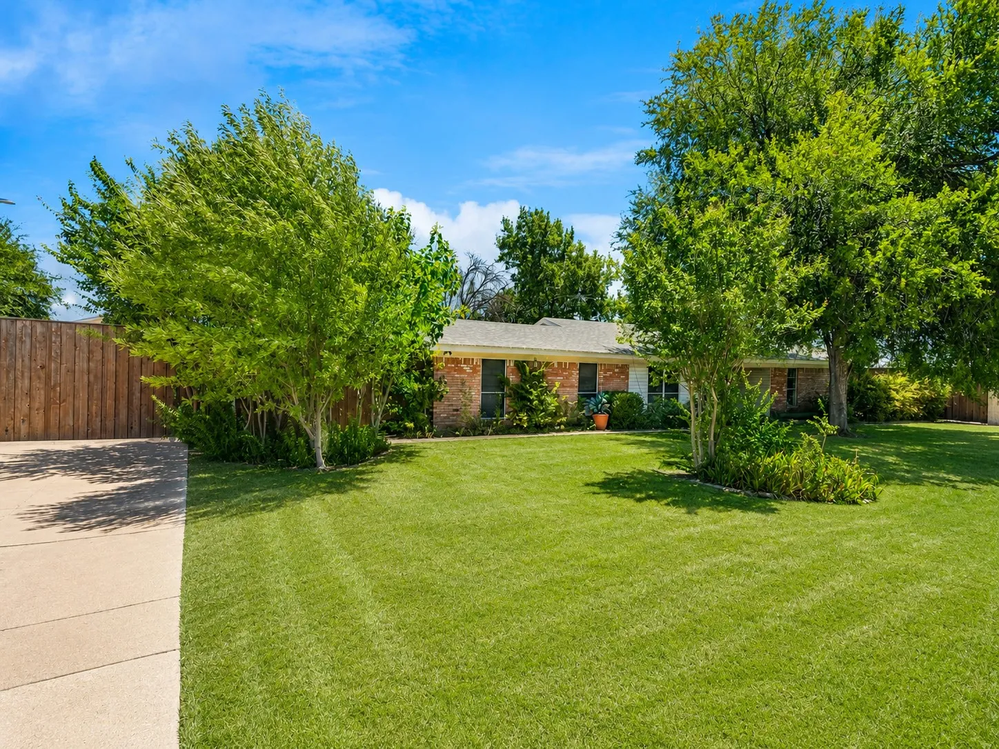 Brick exterior, concrete driveway, mature trees, wood fence, and green lawn