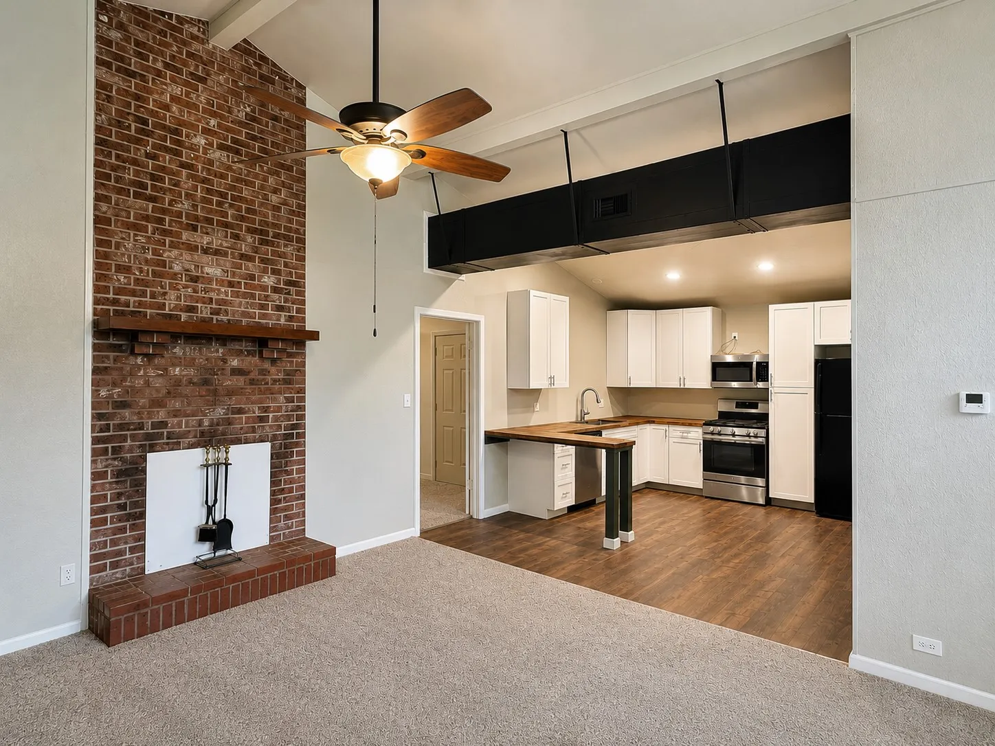 Exposed brick fireplace with a wood mantel, vaulted ceilings, ceiling fan, updated kitchen cabinetry, and wood-finish flooring