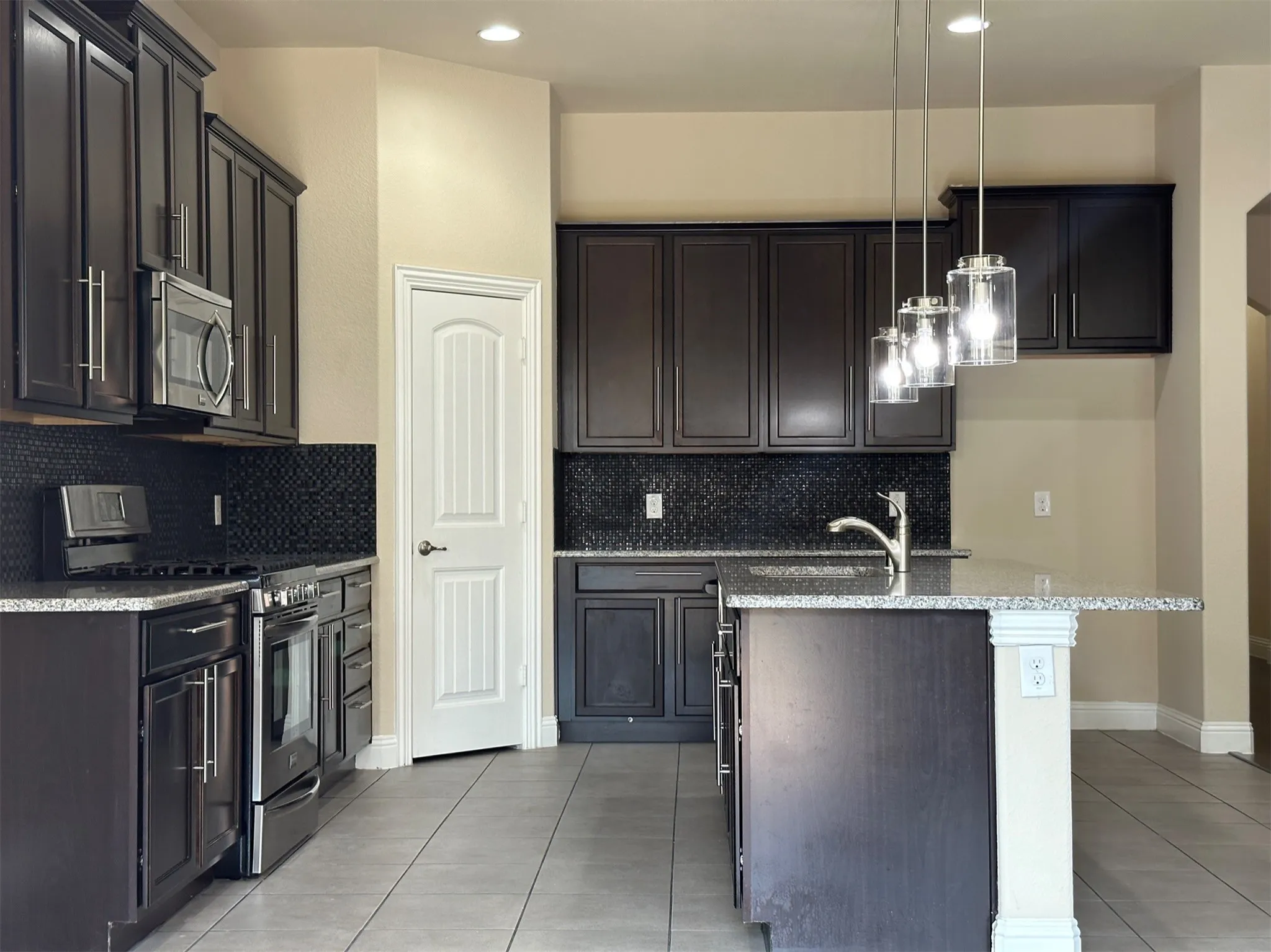 Kitchen featuring dark wood cabinetry, stainless steel appliances, a black mosaic tile backsplash, light-colored countertops, and a central island with pendant lighting