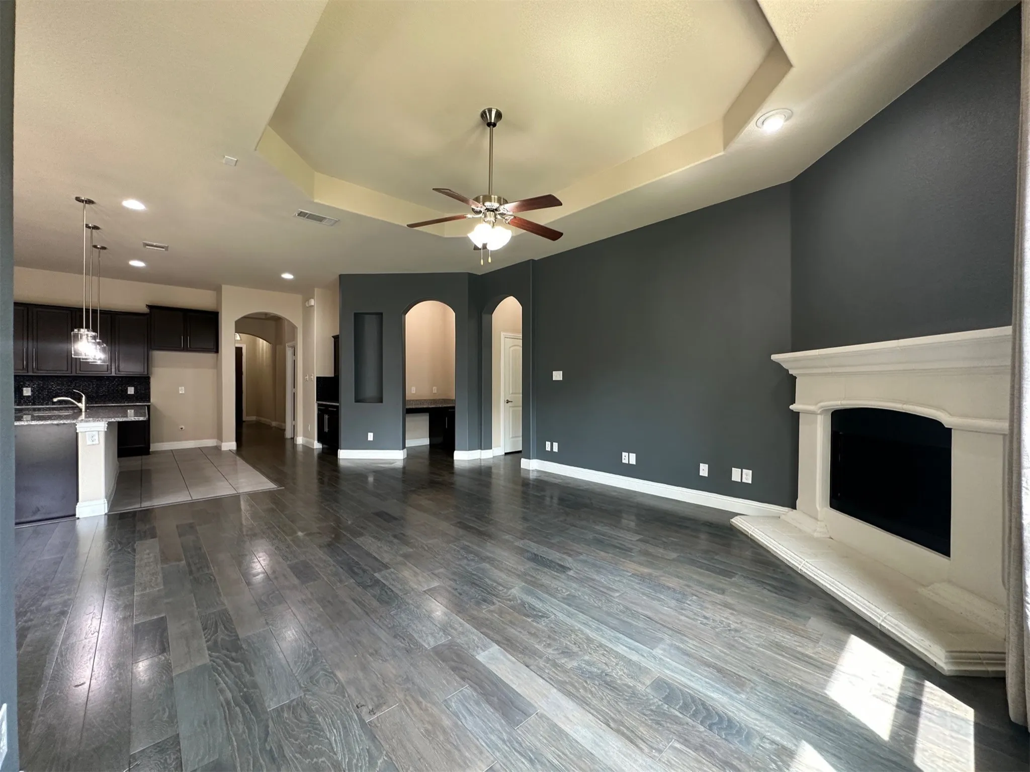 Spacious living area featuring wood-finish flooring, a tray ceiling with a ceiling fan, and a fireplace with a white mantel