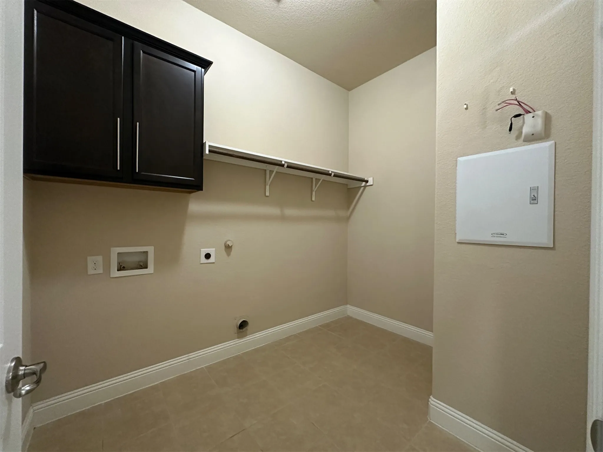 Utility room featuring dark wood-finish cabinetry with silver-tone hardware, a white clothes rod with shelving, light beige ceramic tile flooring, and light beige wall paint with white baseboards