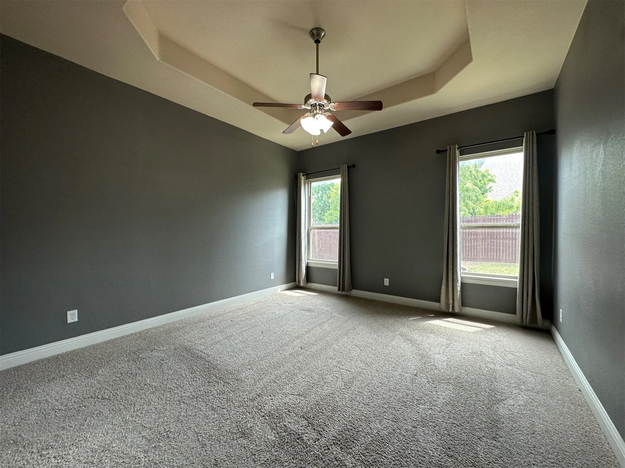 Spacious room featuring a tray ceiling with a ceiling fan, two windows with curtain rods and drapes, neutral-toned wall-to-wall carpeting, and painted baseboards
