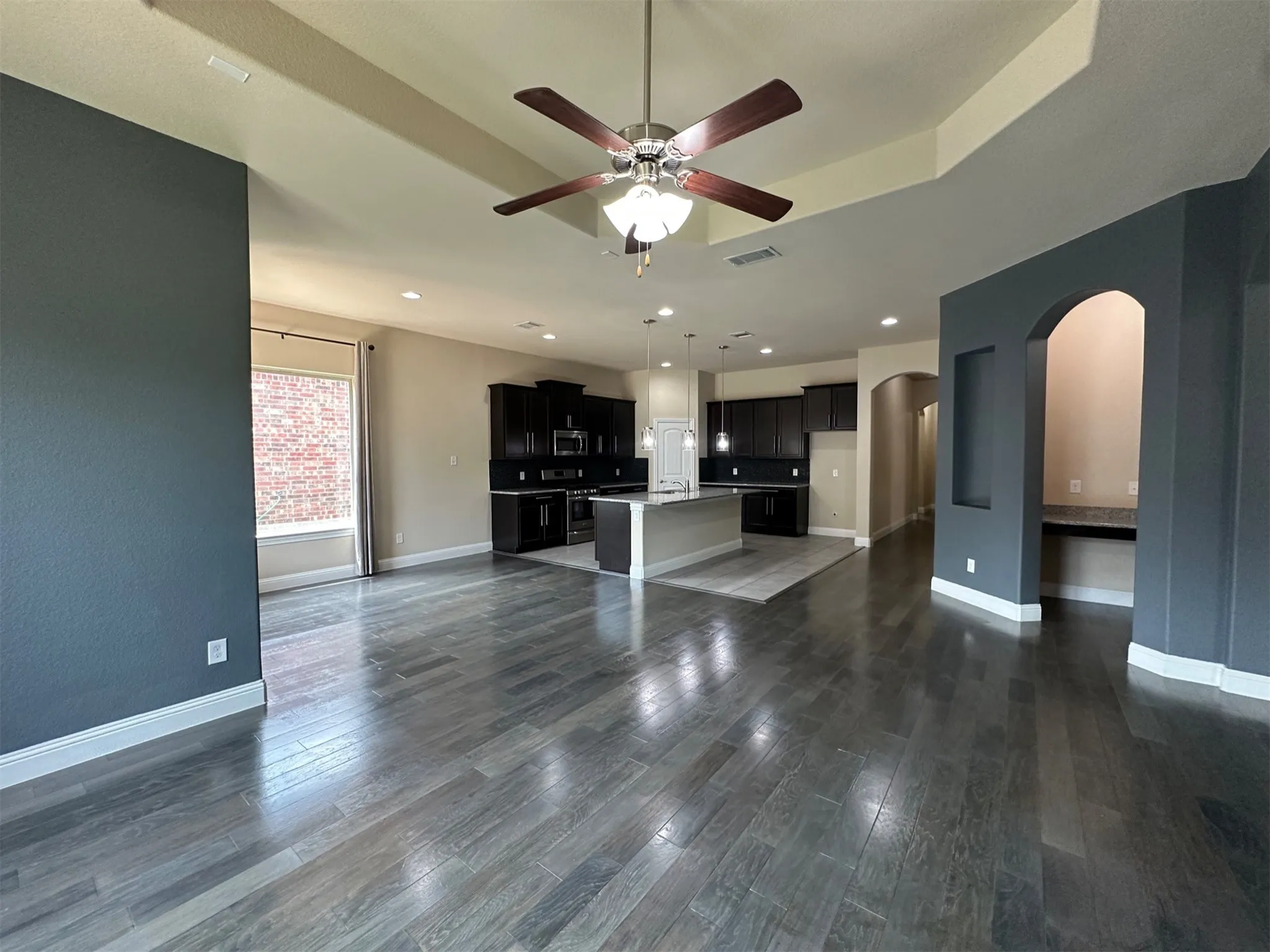 Spacious living area featuring wood-finish flooring, a tray ceiling with a ceiling fan, and a kitchen with dark cabinetry and a central island