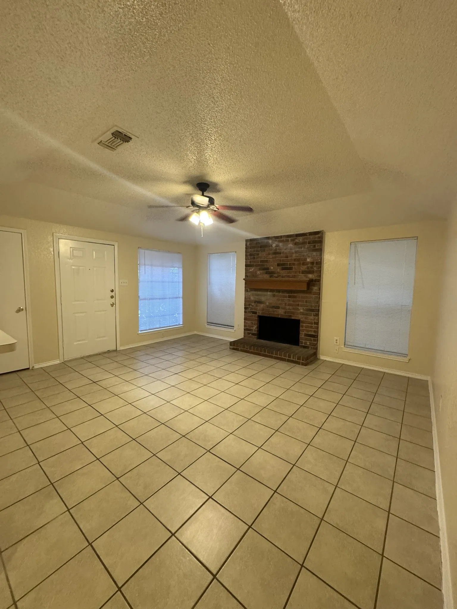 Spacious living area featuring neutral tile flooring, a brick fireplace with a wood mantle, multiple windows, and a ceiling fan