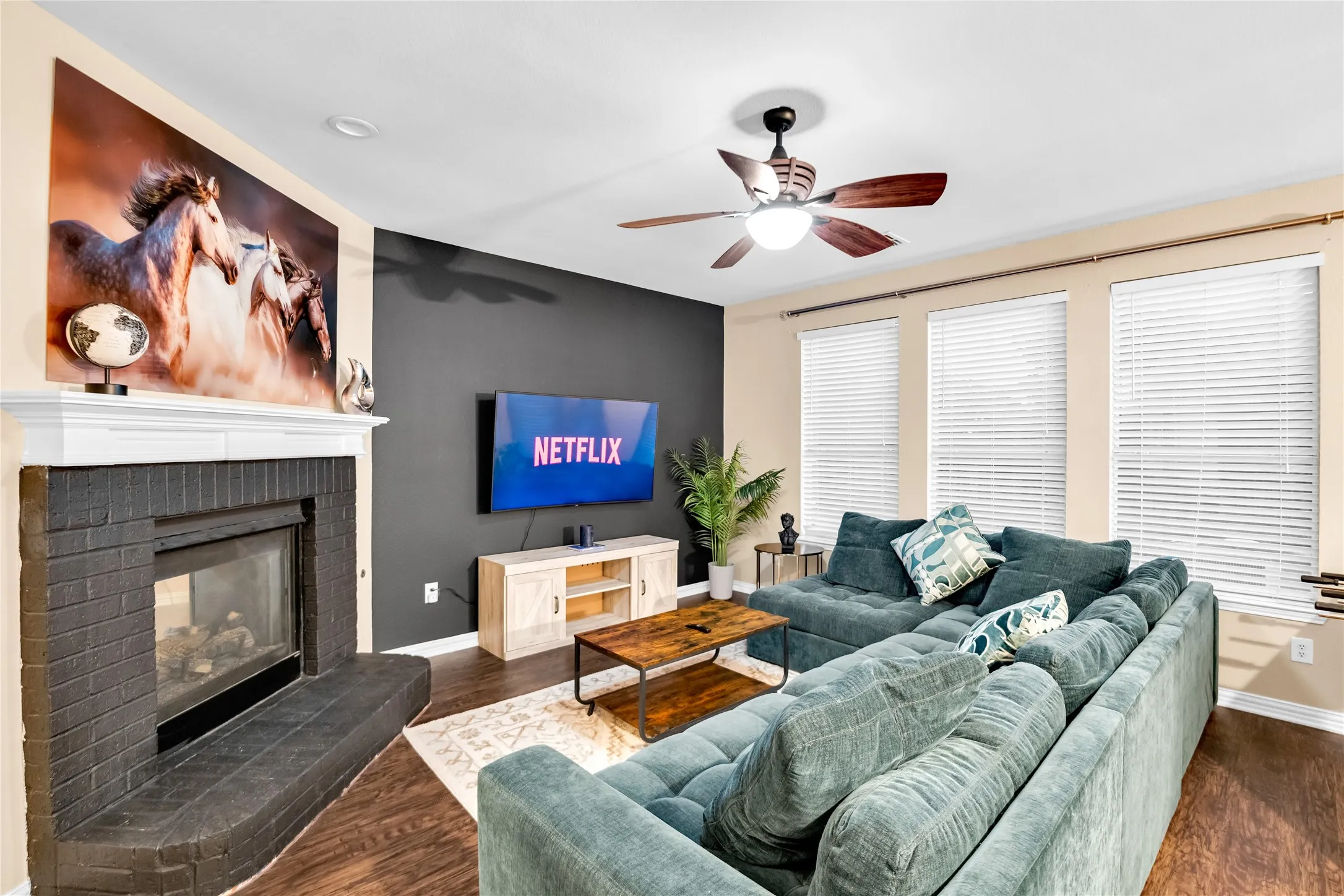 Living area featuring a painted brick fireplace with a white mantel, dark accent wall, wood-finish flooring, and a ceiling fan with integrated lighting